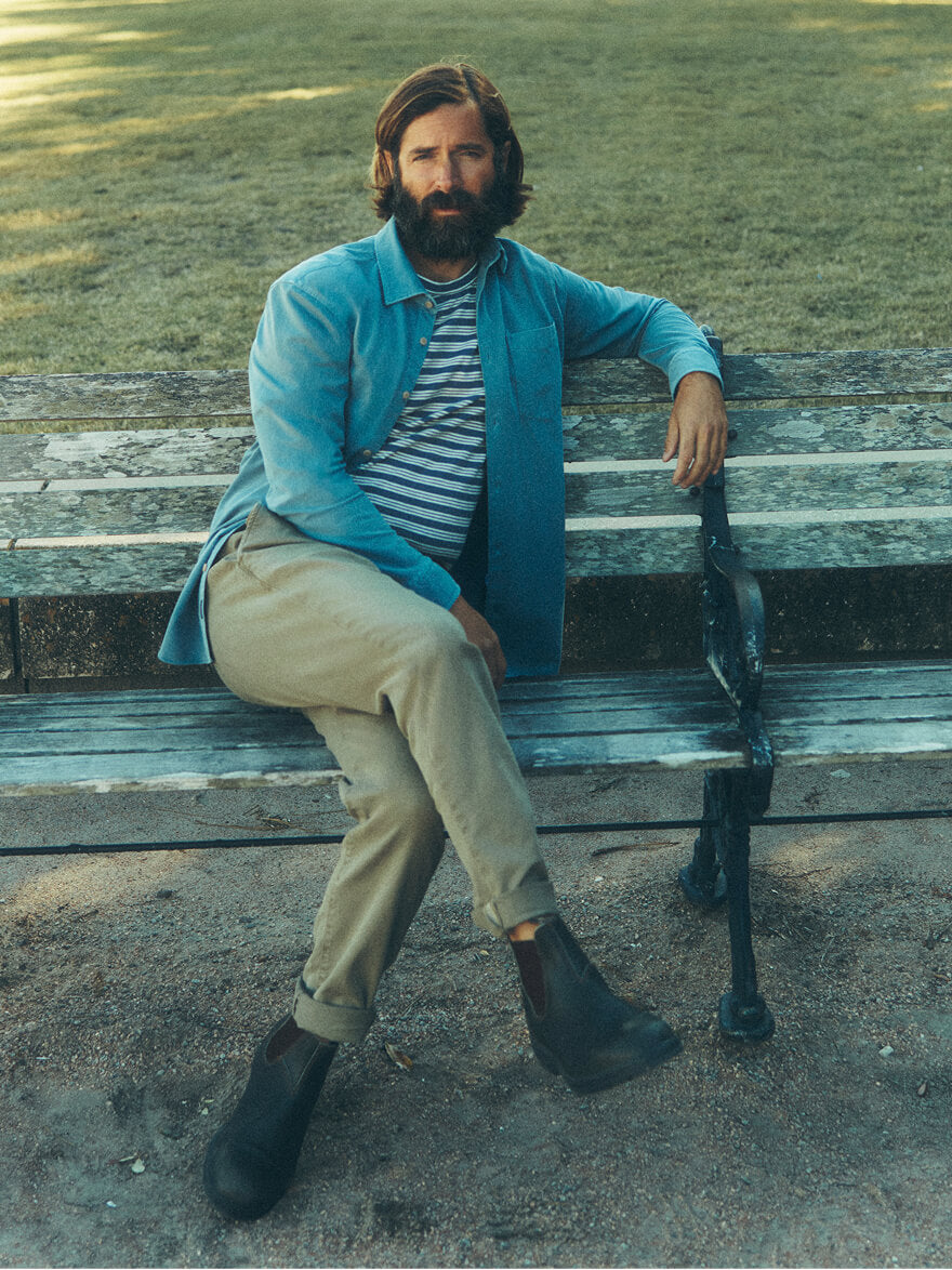 Man seated on a park bench wearing a knit blue shirt over a striped t shirt, stretch terry pants and blundstone boots