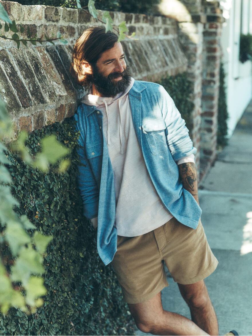 Man leaning on an ivy and brick wall wearing a legend sweater shirt over a slub hoodie paired with corduroy shorts