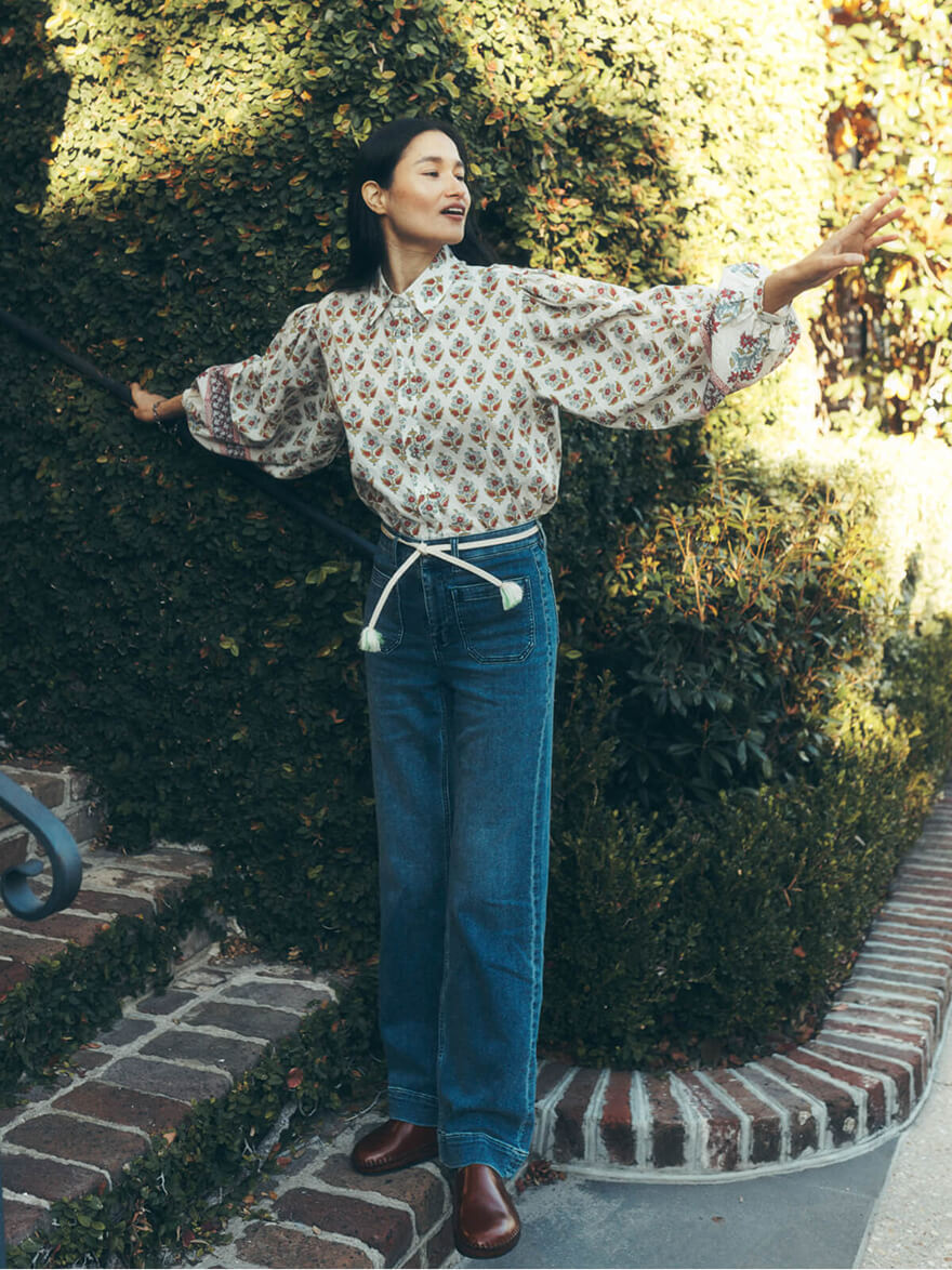 A woman poses next to a hedge wearing a floral print blouse, stretch terry patch pocket pants styled with a rope belt and clogs