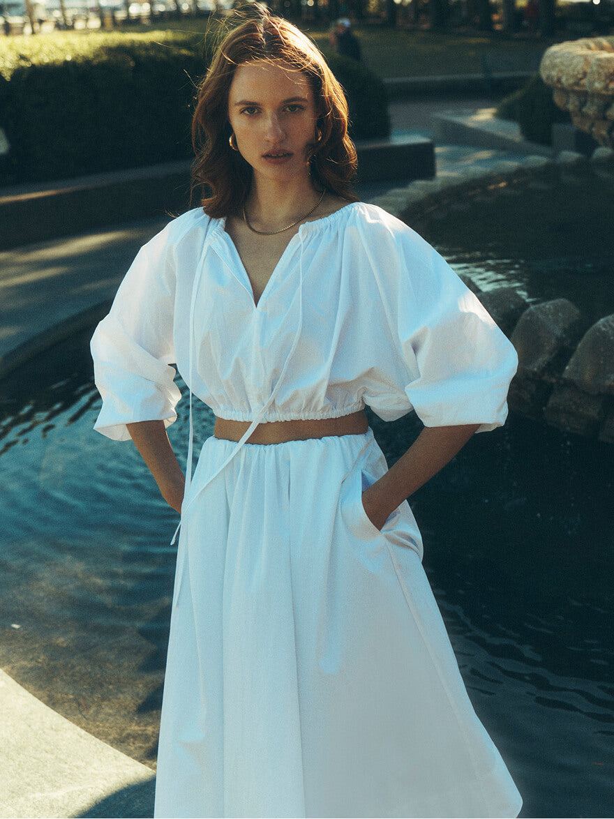 Woman in a white blouse and matching skirt standing in front of a fountain at sunset