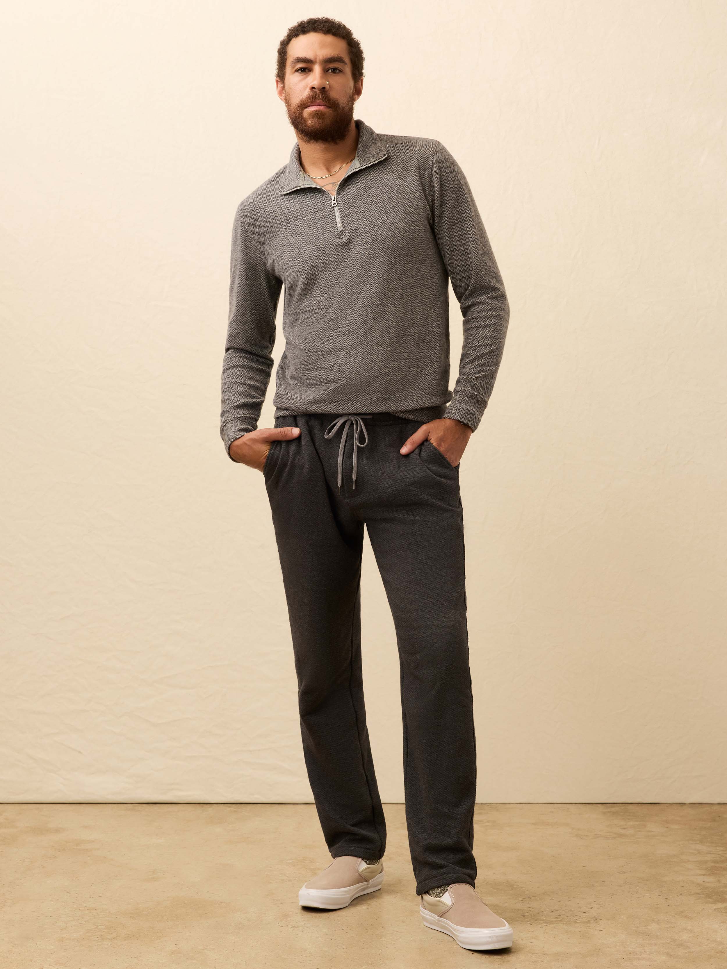 Man wearing a light gray sweater and dark grey jogger pants against a beige background
