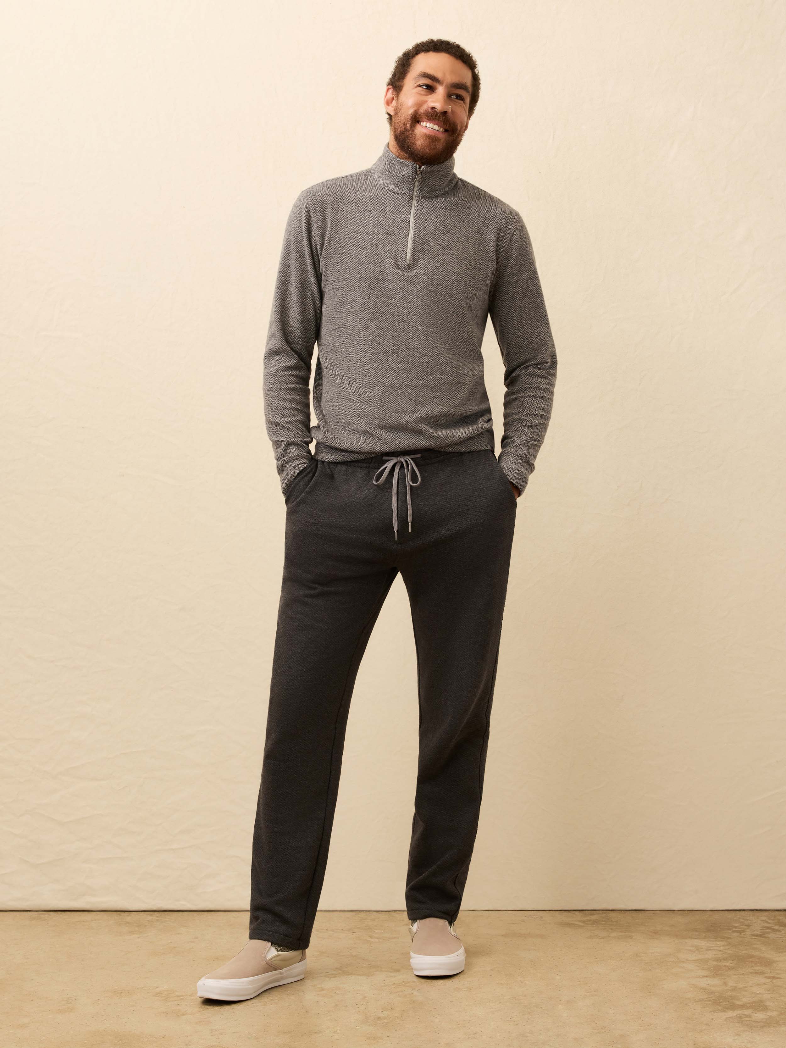Man wearing a light gray sweater and dark grey jogger pants against a beige background