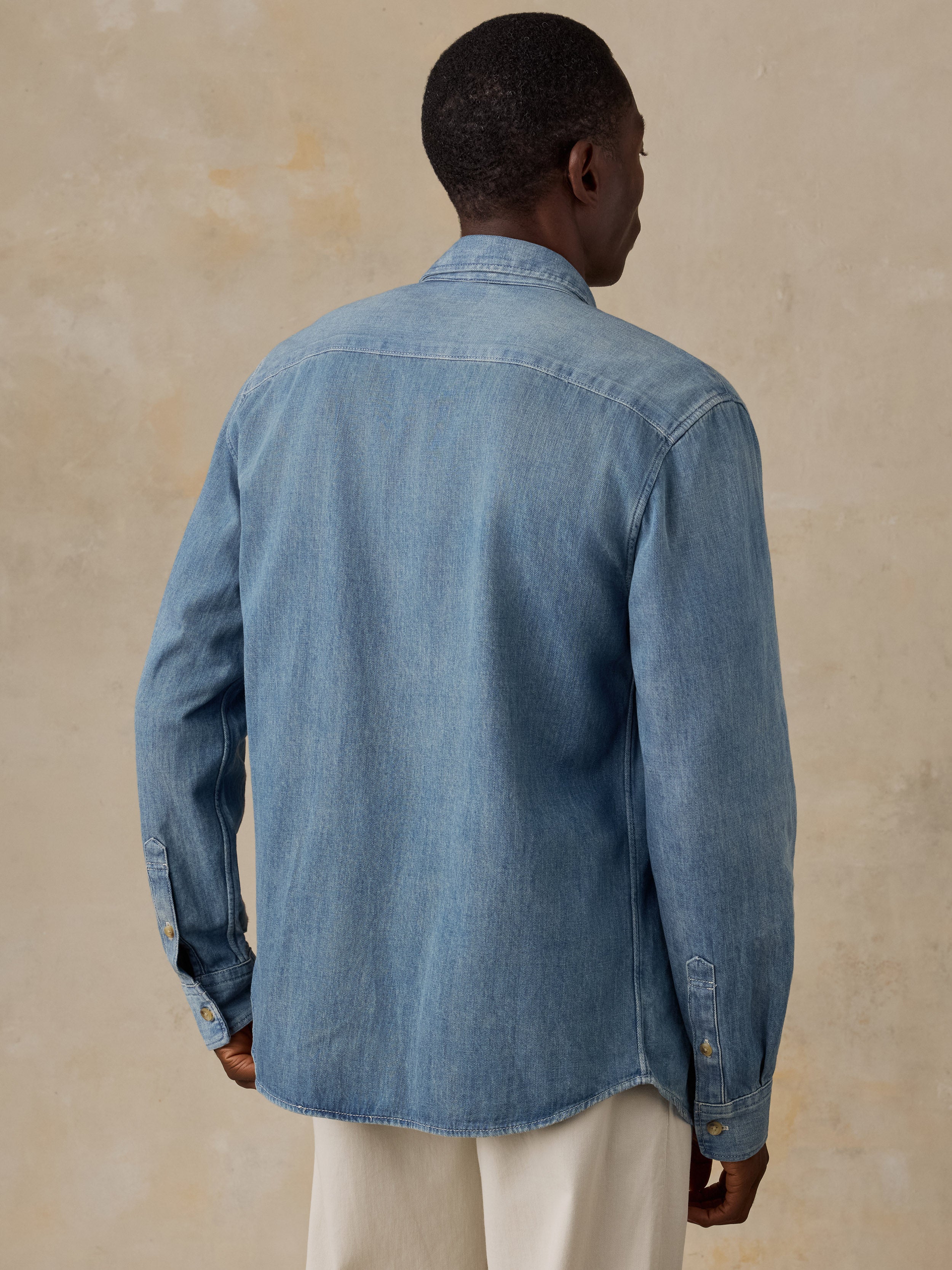 A man viewed from the back wearing a light blue denim long-sleeve button-up shirt and matching blue jeans, standing against a plain beige wall.