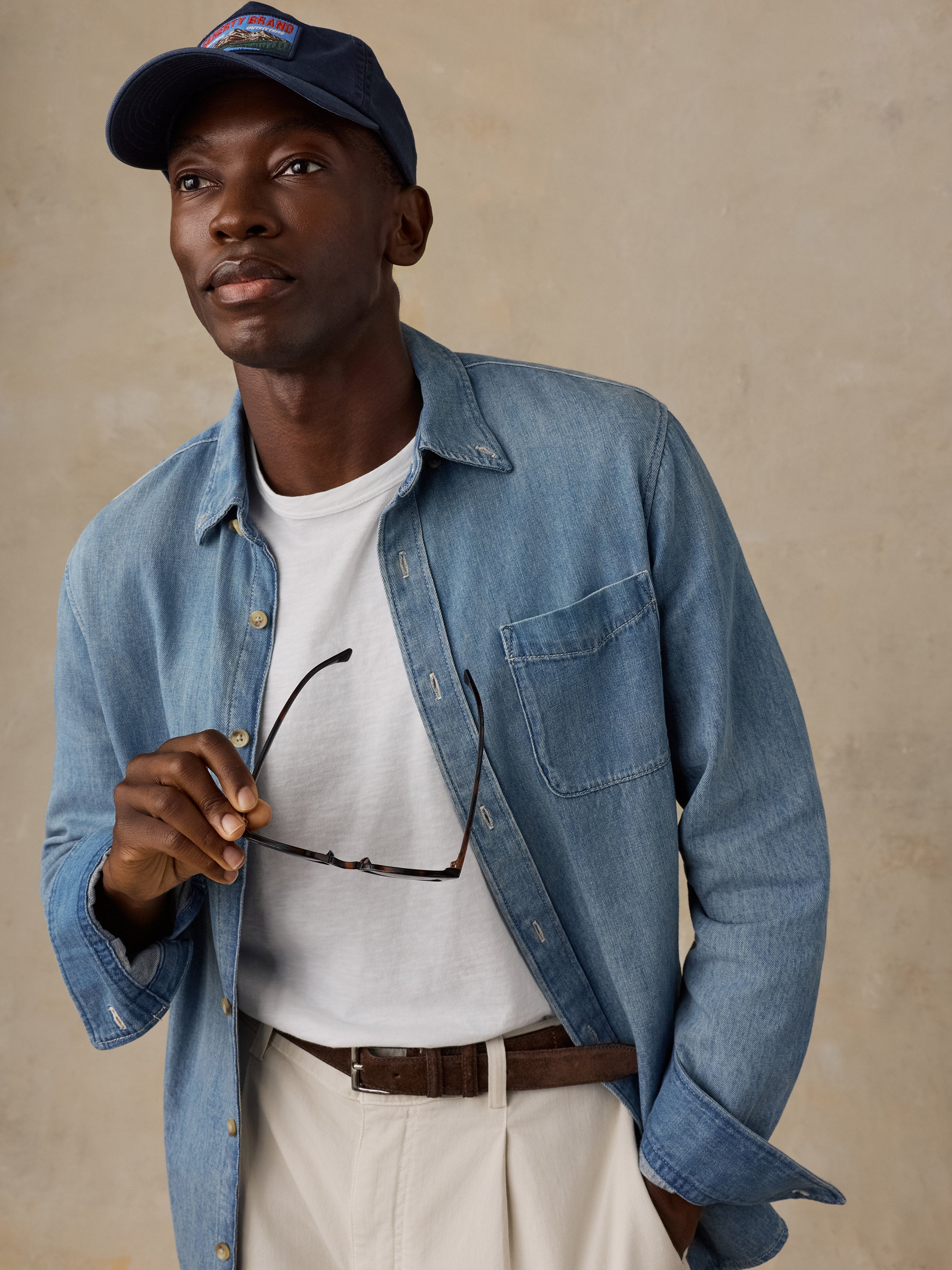 Man wearing a light blue denim shirt over a white t-shirt, khaki pants, and a hat against a beige background
