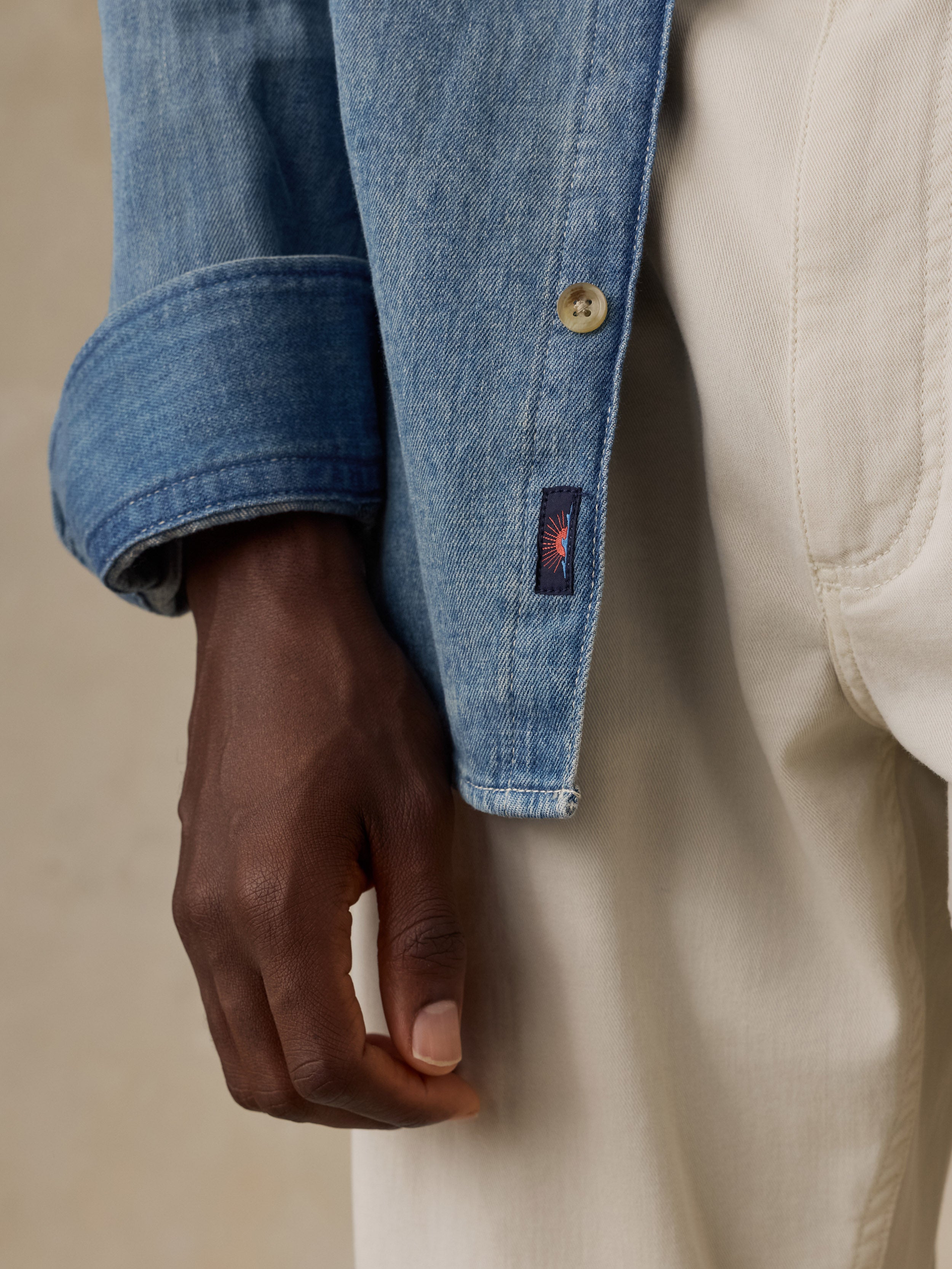 Close-up of a light blue denim button-up shirt showing the button closure, fabric texture, and a small Faherty brand sun and waves patch near the hem