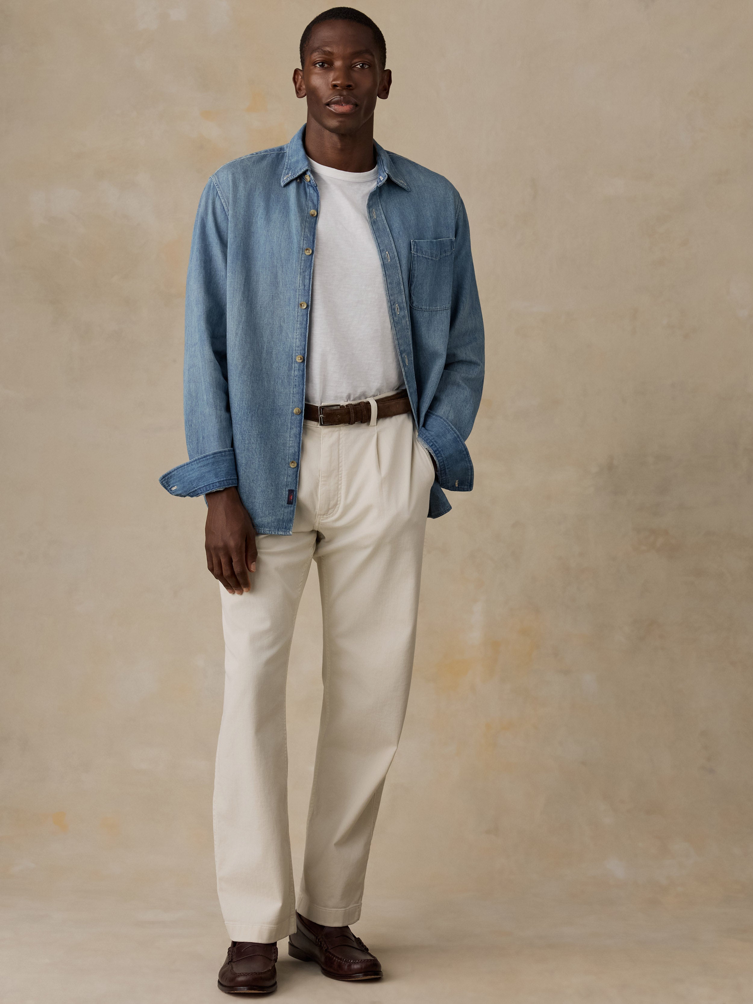 A man wearing a light blue denim shirt and matching light blue jeans, styled with a brown belt and brown loafers, standing against a textured beige wall.