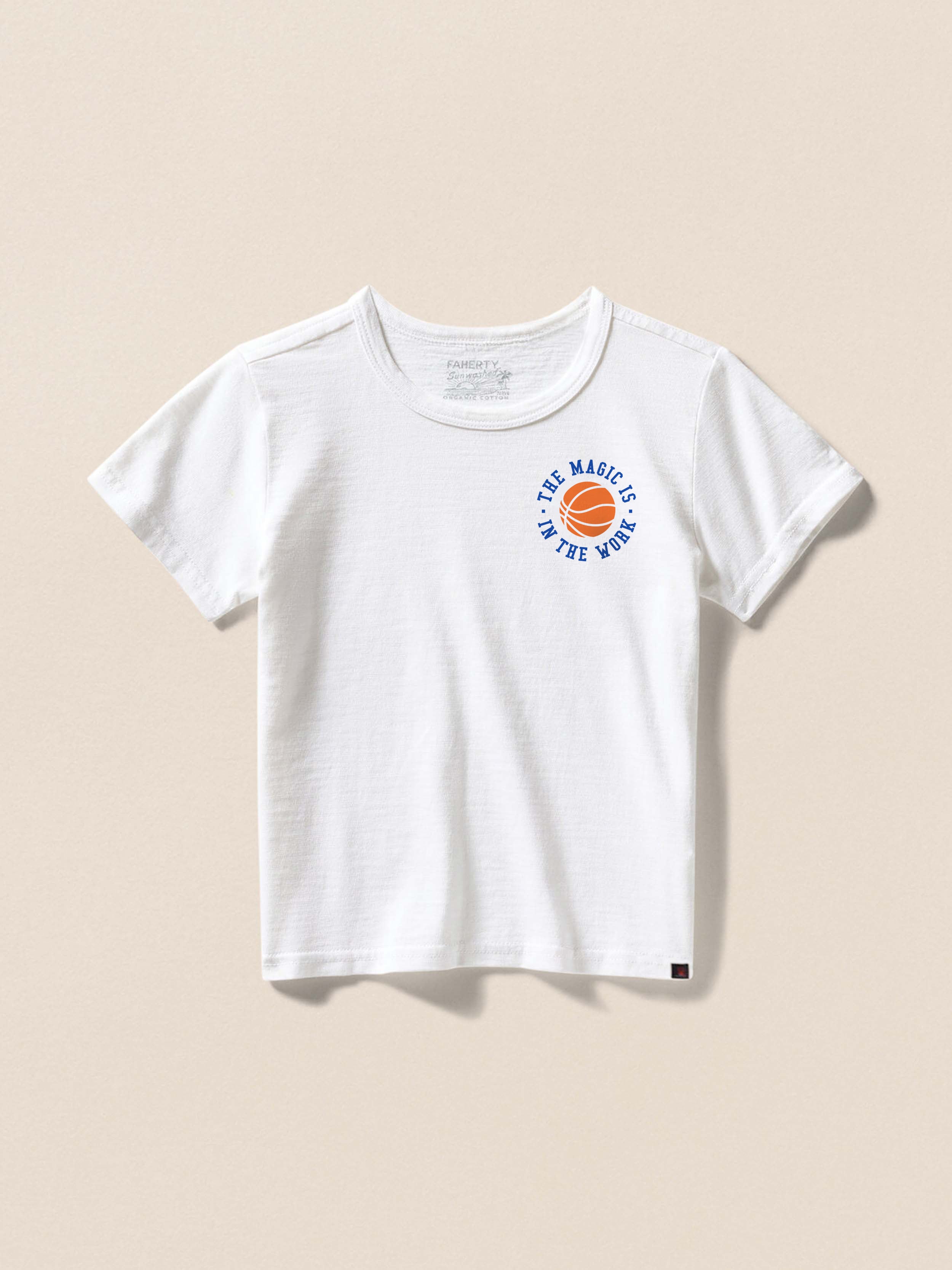 White t-shirt with "The Magic Is In The Work" graphic on the left chest against a beige background