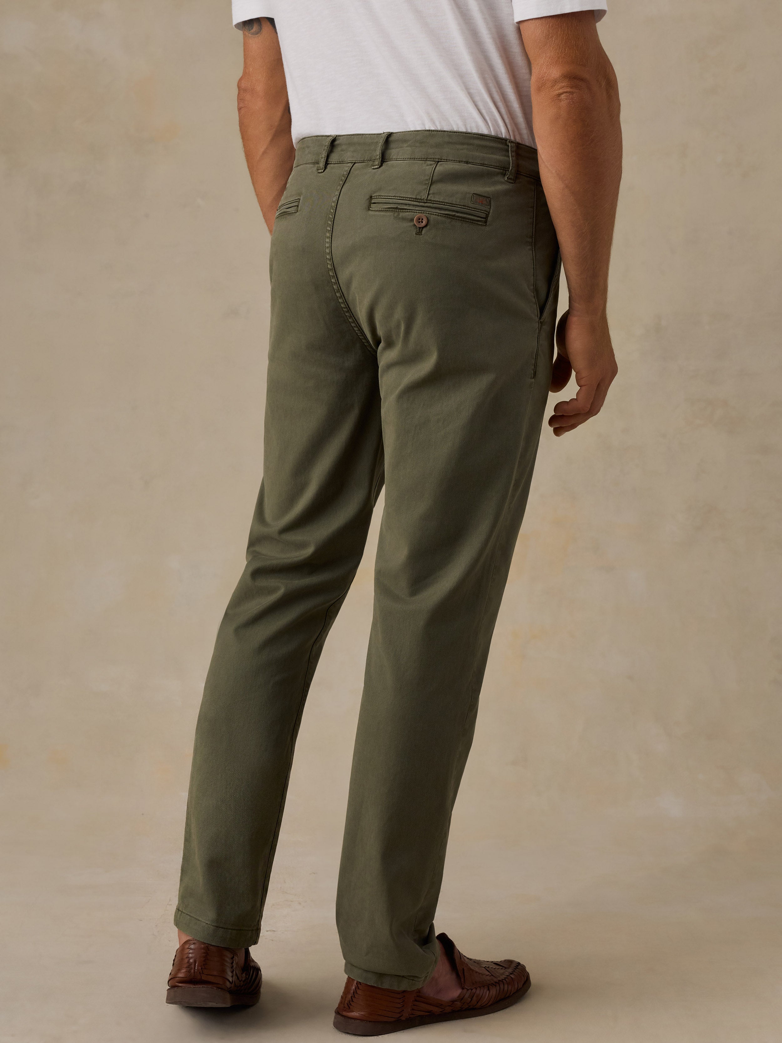 A man wearing olive green chino pants, a white t-shirt, and brown leather loafers, shown from a rear three-quarter angle against a textured beige studio backdrop.
