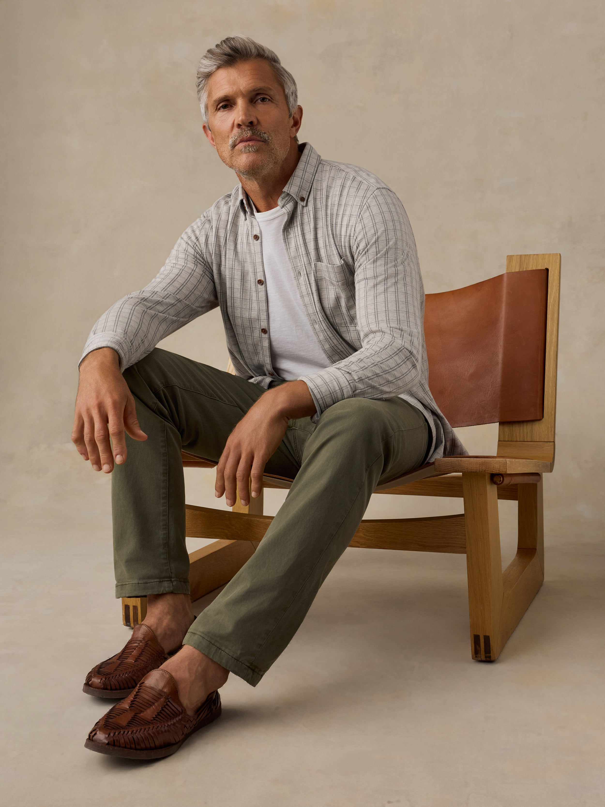 A man sits on a wooden chair with brown leather backrests, wearing a light gray and white striped button-down shirt over a white undershirt, olive green pants, and brown leather loafers, against a textured beige studio backdrop.
