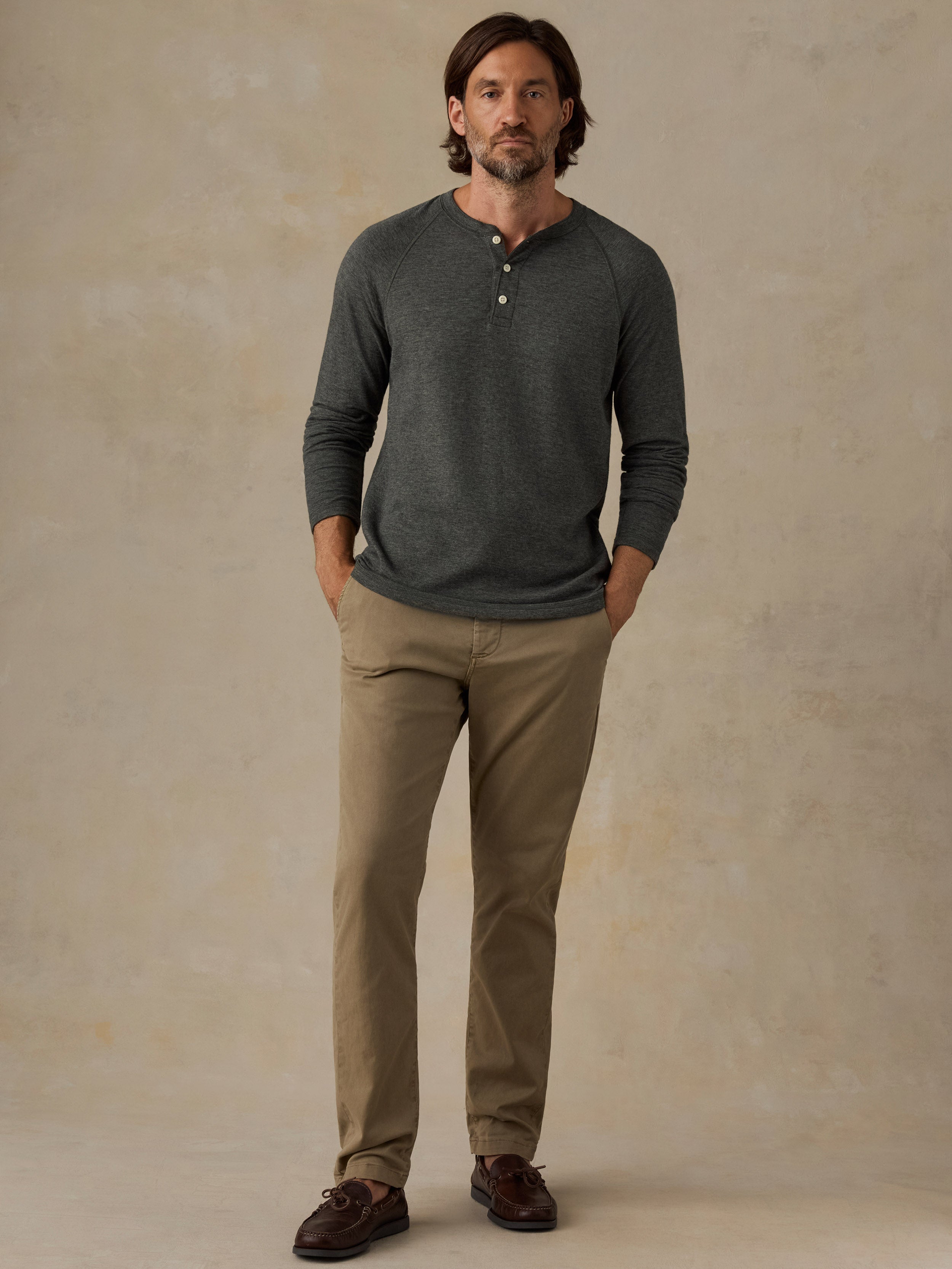 A man wearing a dark gray long-sleeve henley shirt, khaki pants, and brown shoes, standing against a textured beige studio backdrop.