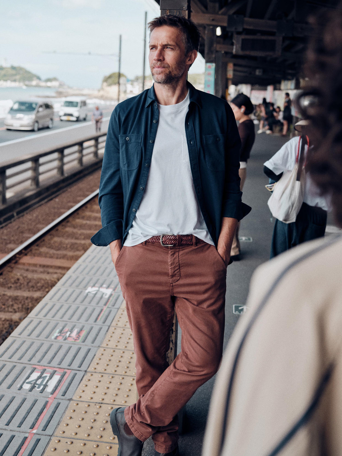A man standing on a train station platform wearing a dark navy overshirt over a white t-shirt, brown trousers, and dark shoes. The background shows a blurred train station environment with tracks, cars, and other passengers., image 1 of 7