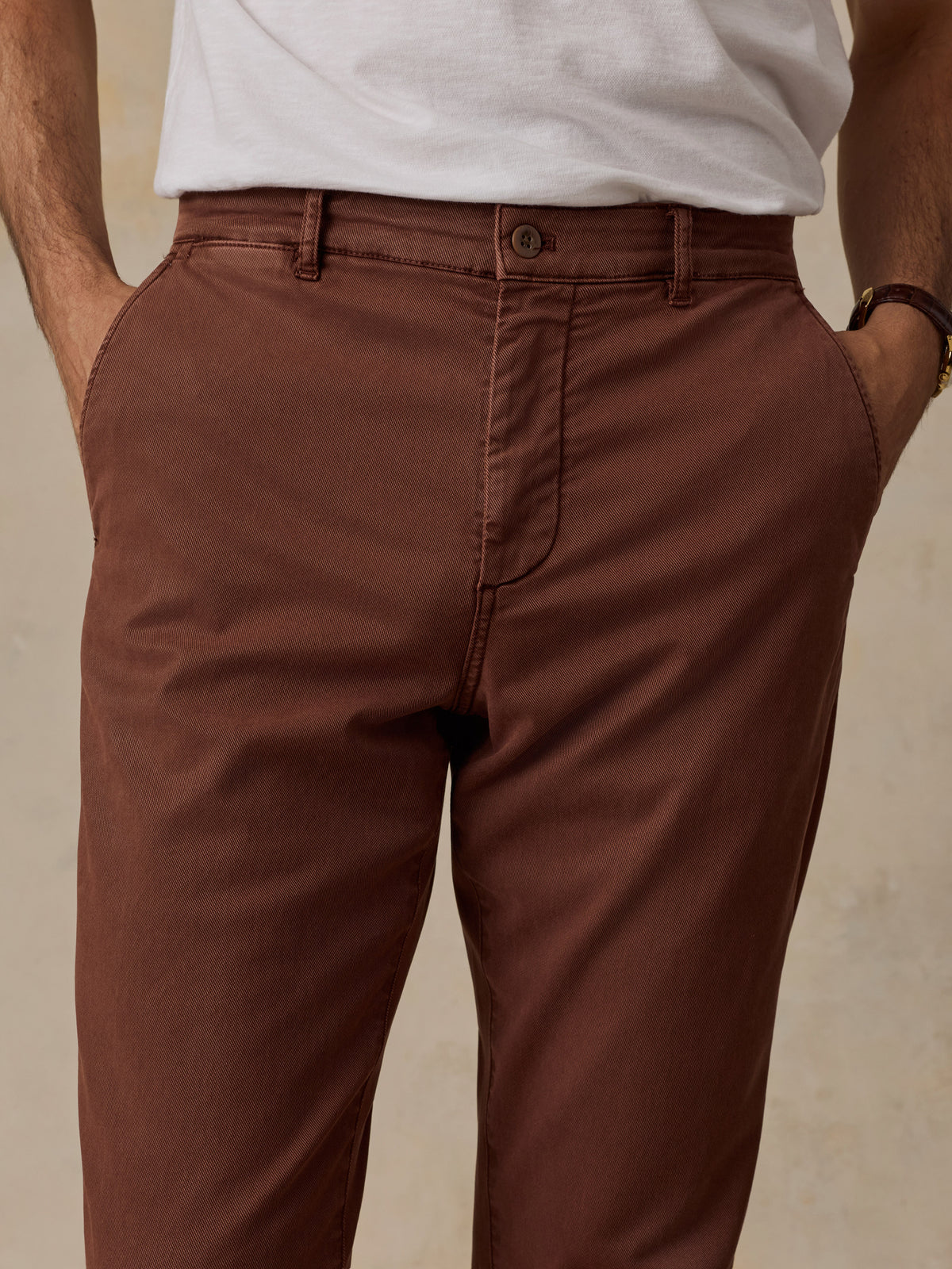 A close-up of a person wearing brown chino pants with a white t-shirt, standing with hands in pockets against a neutral beige background., image 6 of 7