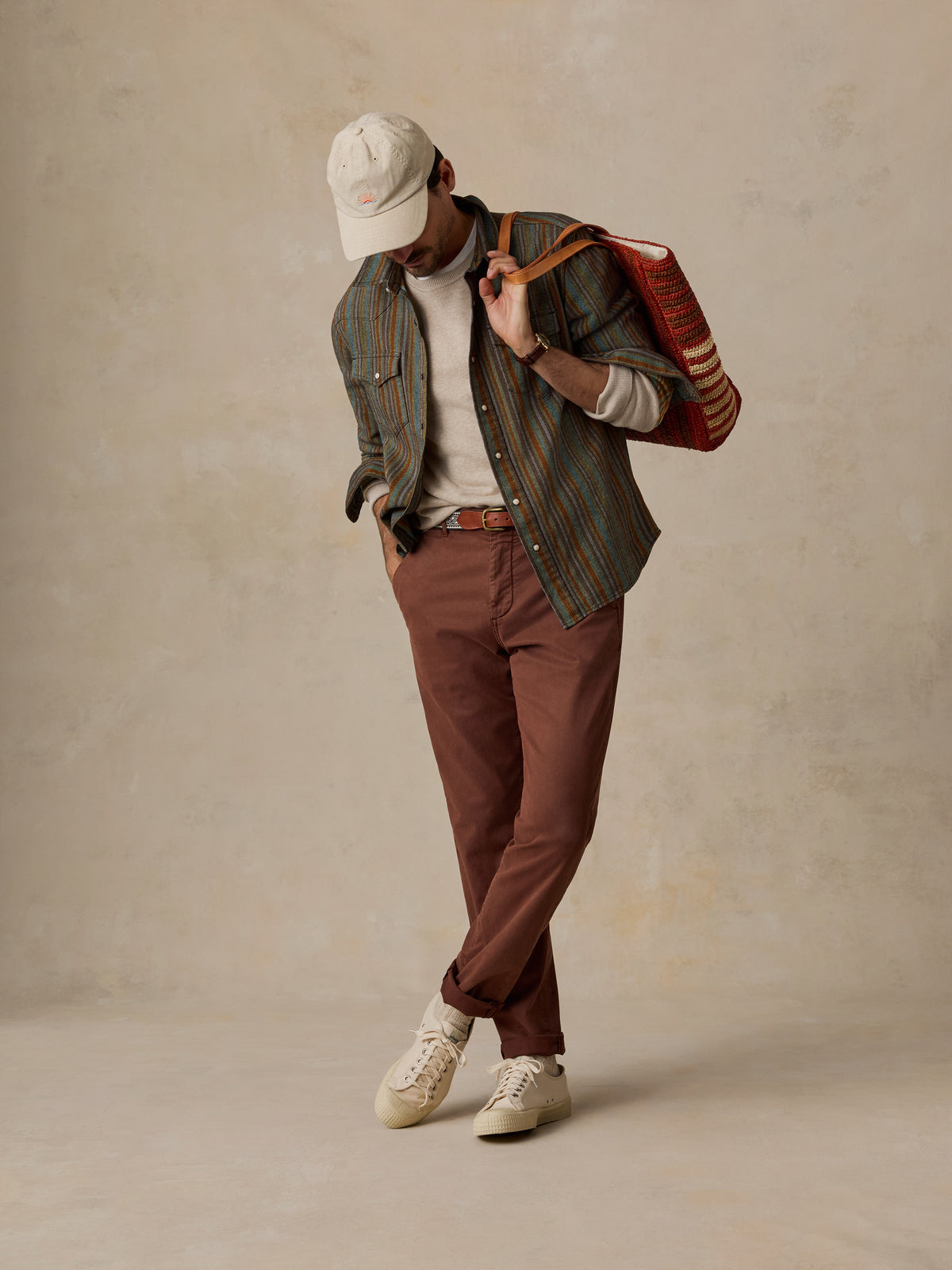 A person wearing a striped button-up shirt, brown trousers, a white cap, and white sneakers, carrying a red and brown striped tote bag against a textured beige studio backdrop., image 3 of 7