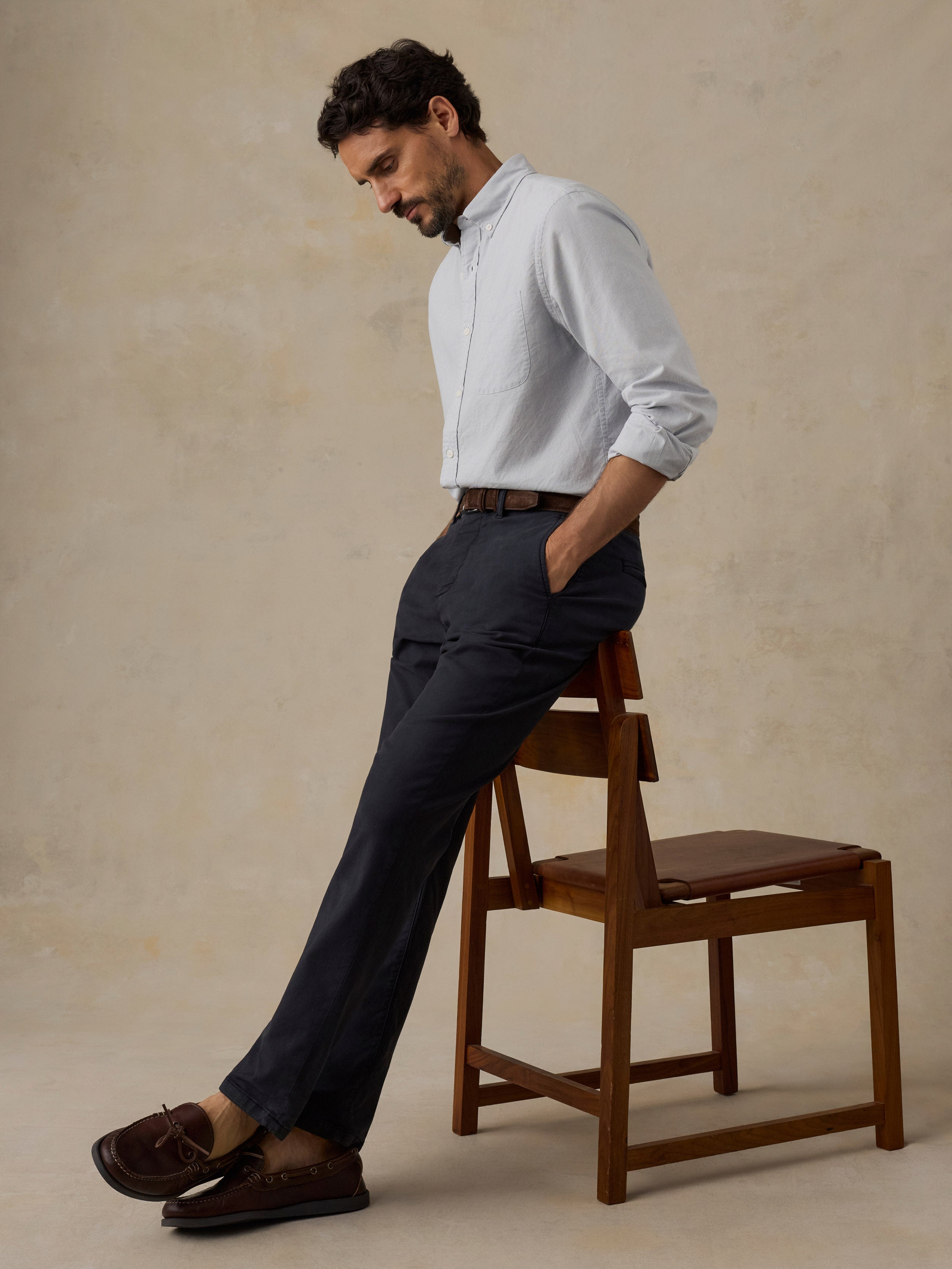 A man wearing a light blue long-sleeve button-down shirt, black trousers, and brown loafers leans against a wooden chair in a studio setting with a textured beige background.