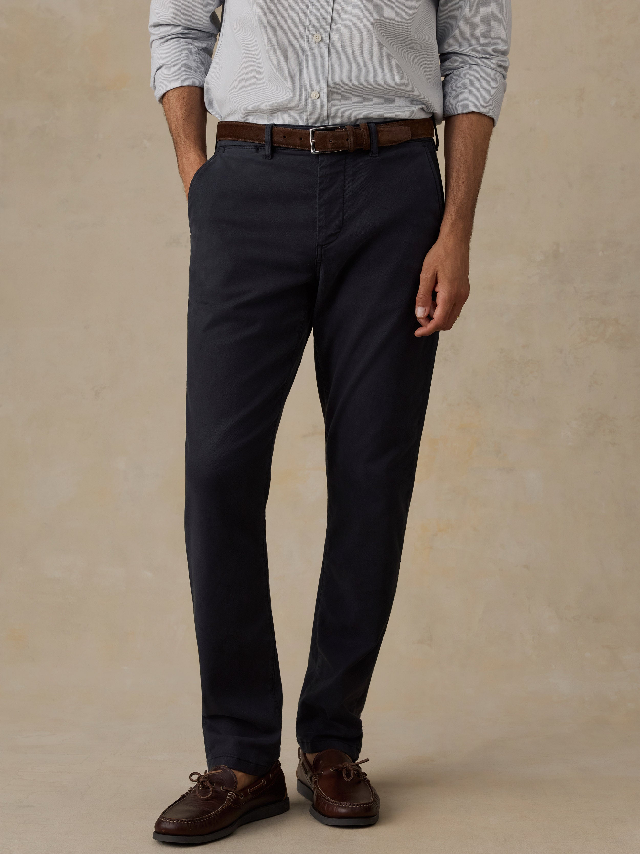 A man wearing black trousers, a light gray button-down shirt, a brown belt, and brown boat shoes, standing against a textured beige studio backdrop.
