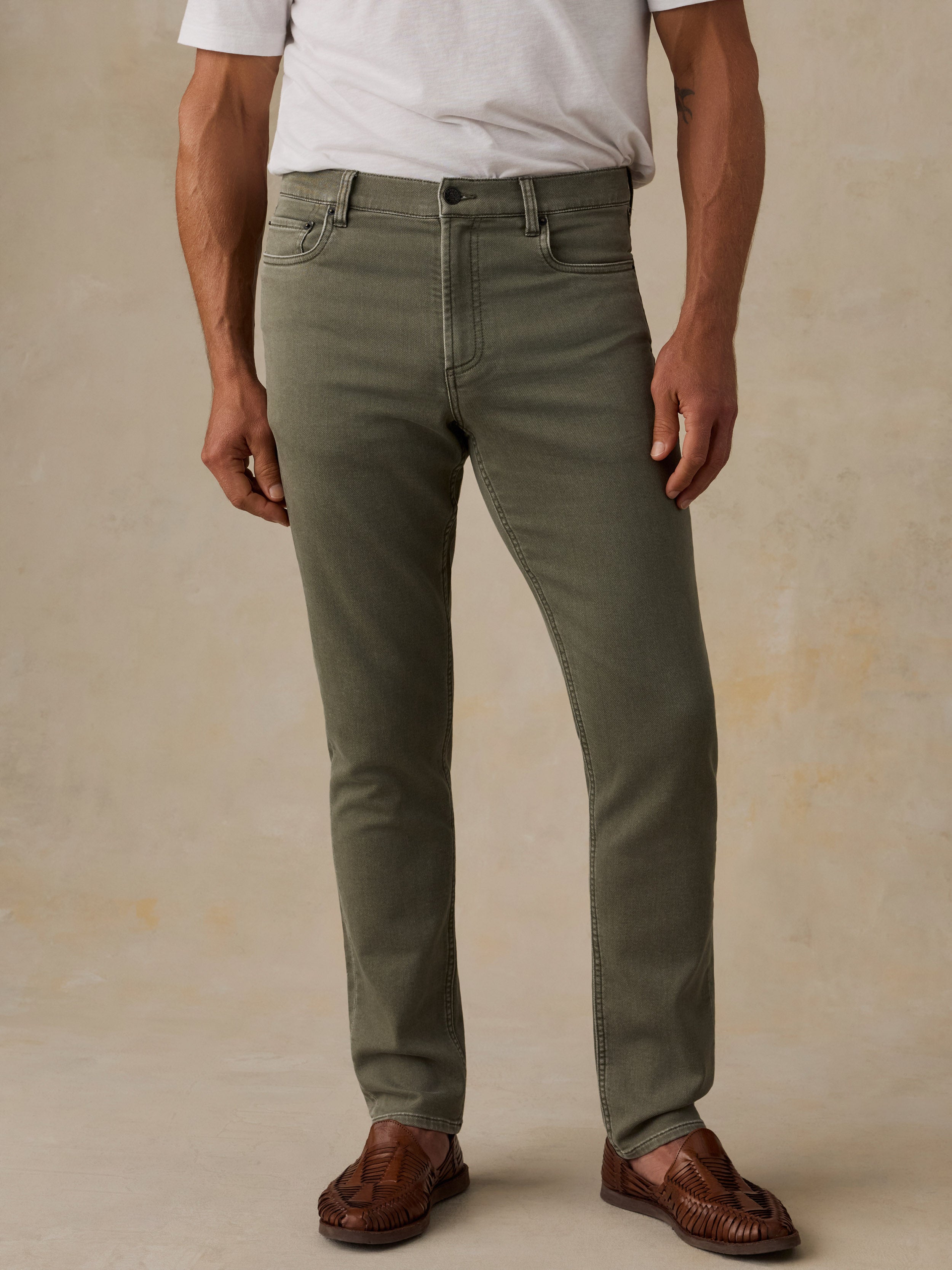 A man wearing slim-fit olive green jeans, a white t-shirt, and brown leather loafers, standing against a textured beige studio backdrop.