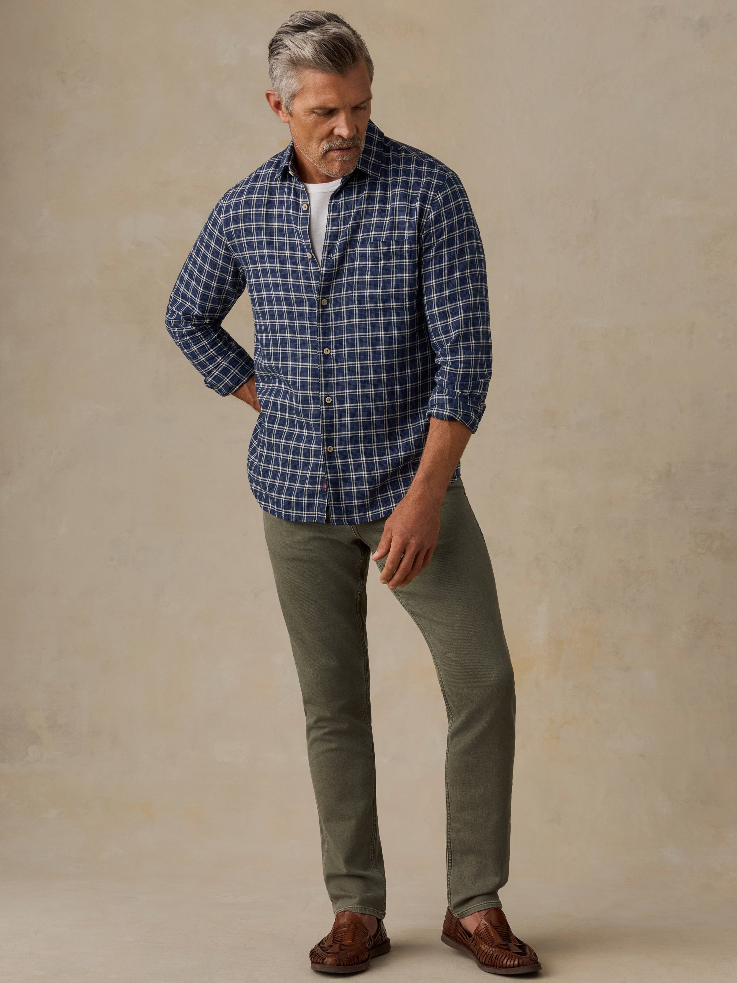 A man wearing a blue and white plaid button-down shirt, olive green pants, and brown leather loafers, standing against a textured beige studio backdrop.