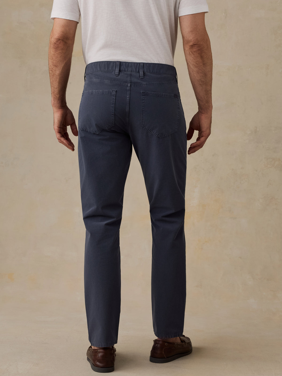 Rear view of a man wearing navy blue slim-fit trousers, a white t-shirt, and brown leather loafers, standing against a textured beige studio backdrop., image 3 of 5
