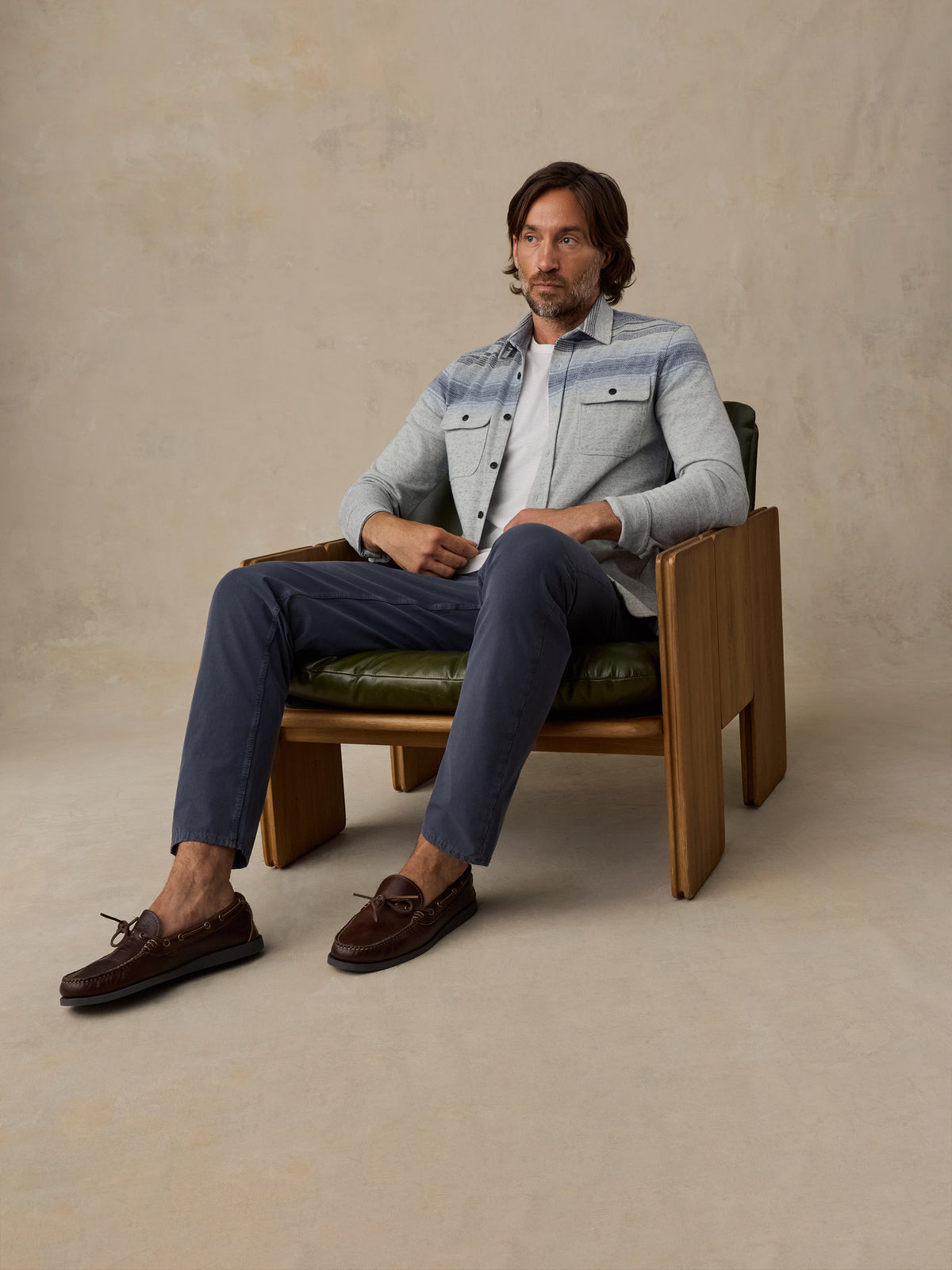 A man sits in a wooden armchair wearing a light blue denim jacket over a white shirt, navy trousers, and dark brown loafers, against a textured beige studio backdrop., image 4 of 5