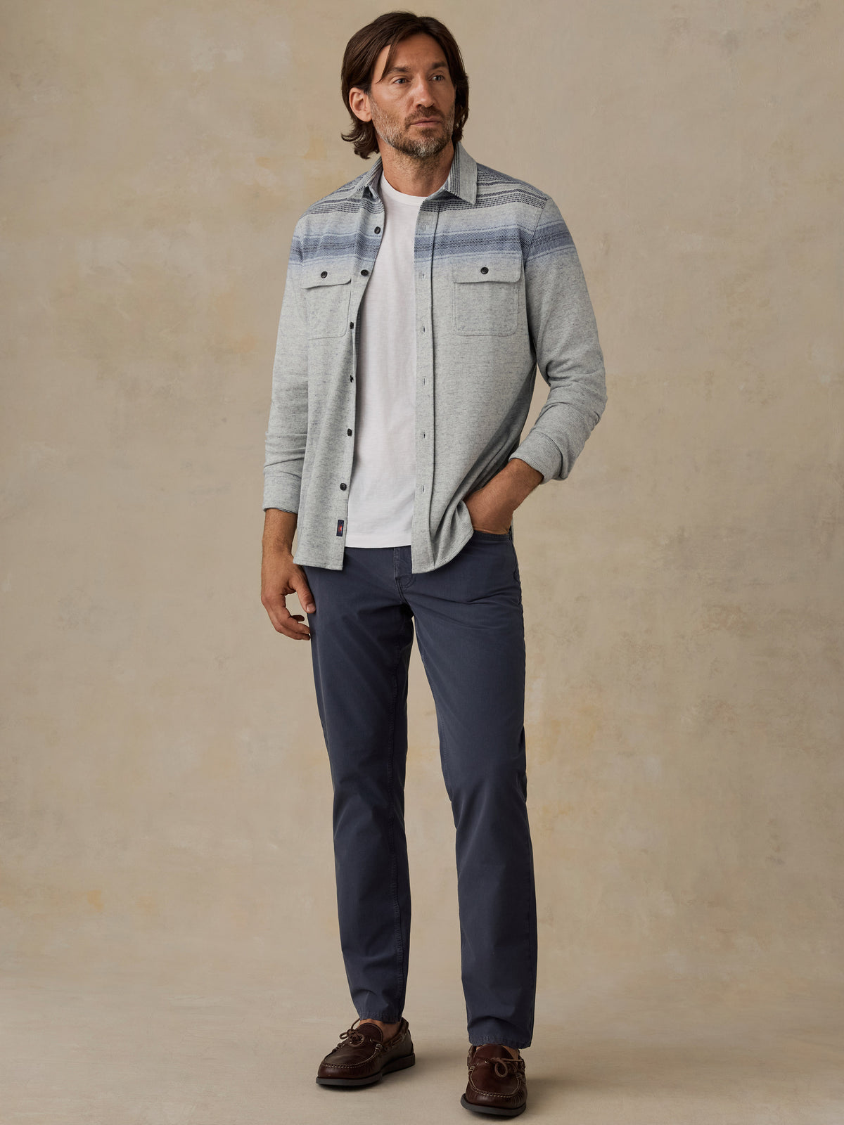 A man wearing a light gray heathered button-down shirt with blue stripes, a white t-shirt, navy blue pants, and brown leather shoes, standing against a textured beige background., image 2 of 5