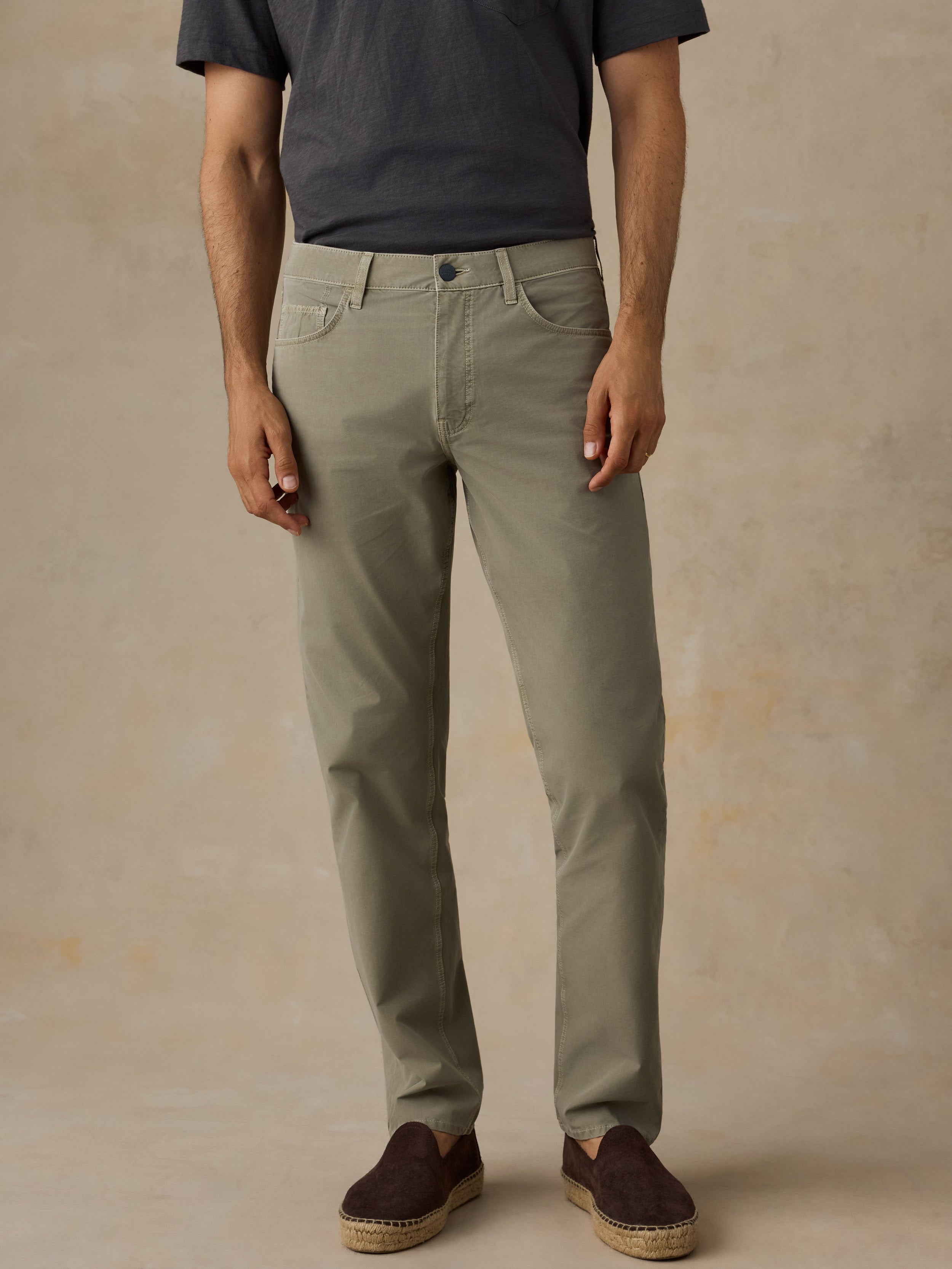 A man wearing slim-fit olive green chinos, a dark charcoal t-shirt, and brown espadrille loafers, standing against a textured beige studio backdrop.