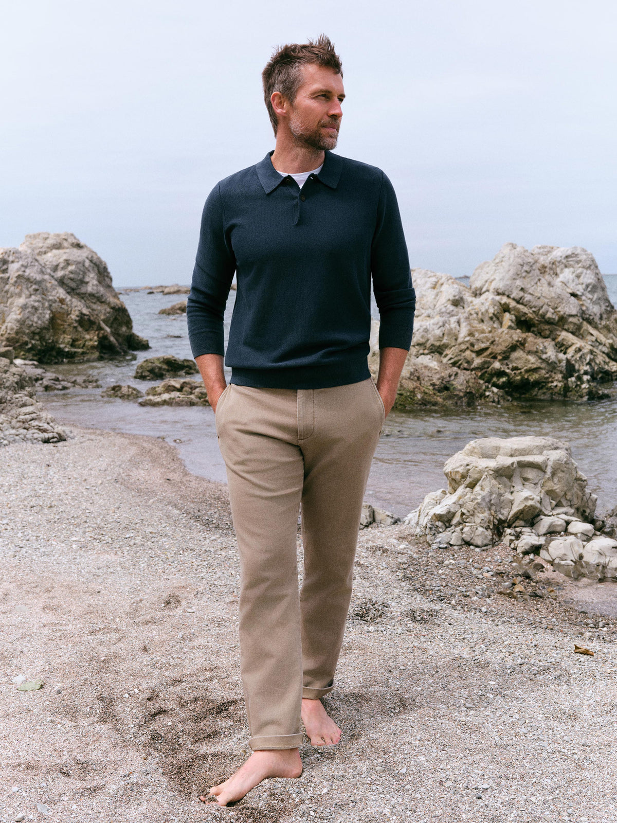 A man standing barefoot on a rocky beach shoreline, wearing a dark navy long-sleeve polo shirt and beige trousers., image 1 of 6