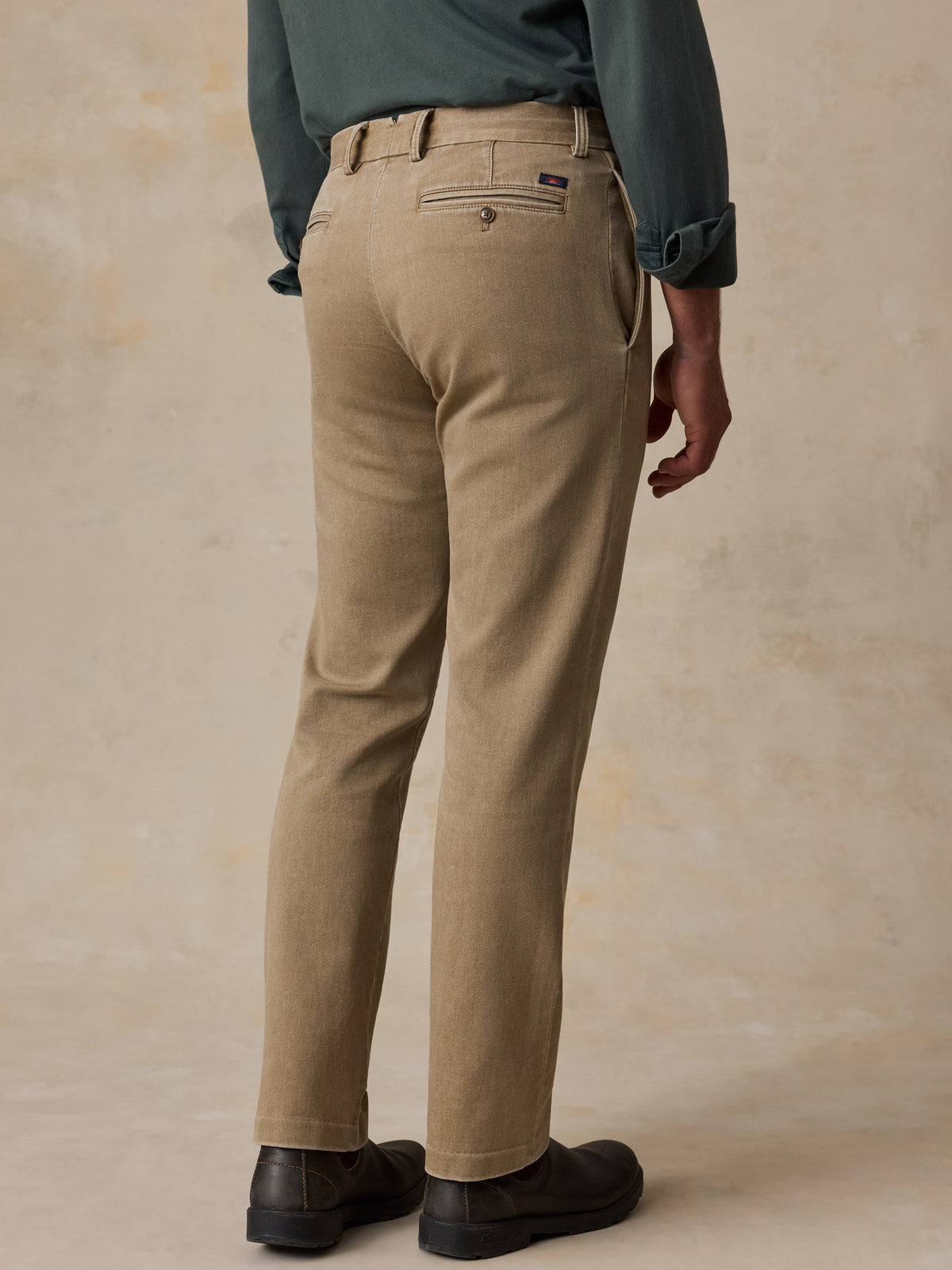 A man shown from the back wearing tan chinos with a dark green shirt and black shoes, standing against a textured beige studio backdrop., image 4 of 6