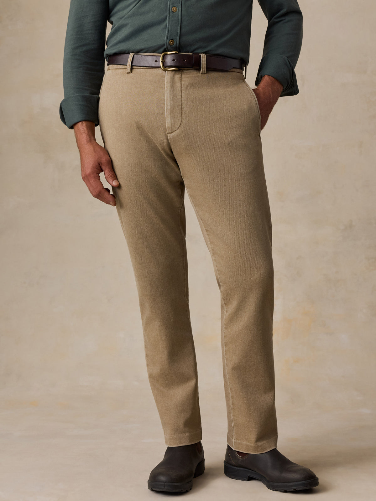 A man wearing khaki chinos with a dark brown belt, a muted green button-down shirt, and black leather boots, standing against a textured beige studio backdrop., image 2 of 6