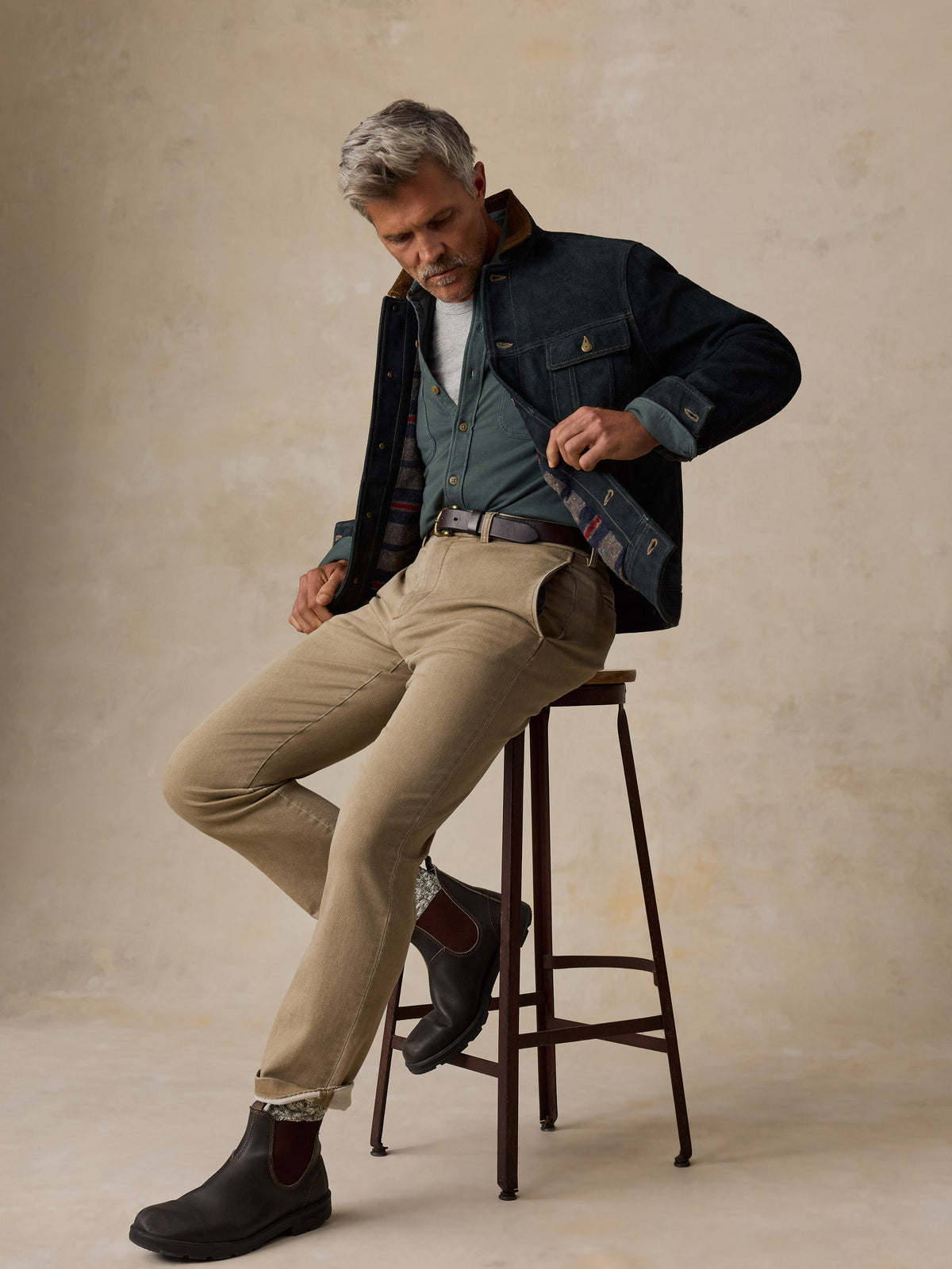 A man with gray hair sits on a dark wooden stool, wearing a layered outfit consisting of a dark navy overshirt, a green button-down shirt, a white t-shirt, beige trousers, and dark brown Chelsea boots, against a textured beige studio backdrop., image 3 of 6