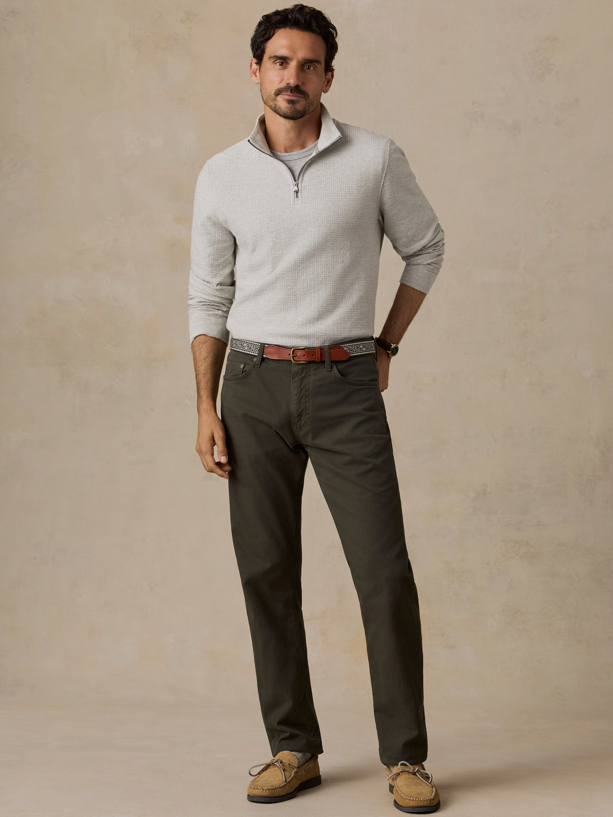 A man modeling a light gray quarter-zip knit sweater, dark olive green trousers, a brown leather belt, and tan boat shoes against a textured beige studio backdrop., image 3 of 6