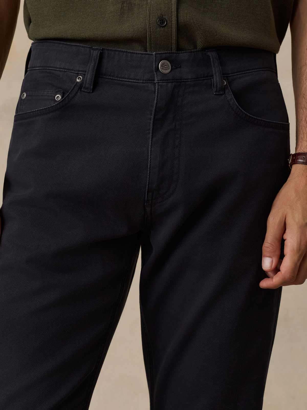 Close-up of a person wearing dark navy blue jeans with a button closure and silver rivets, paired with an olive green shirt, against a neutral beige background., image 5 of 6