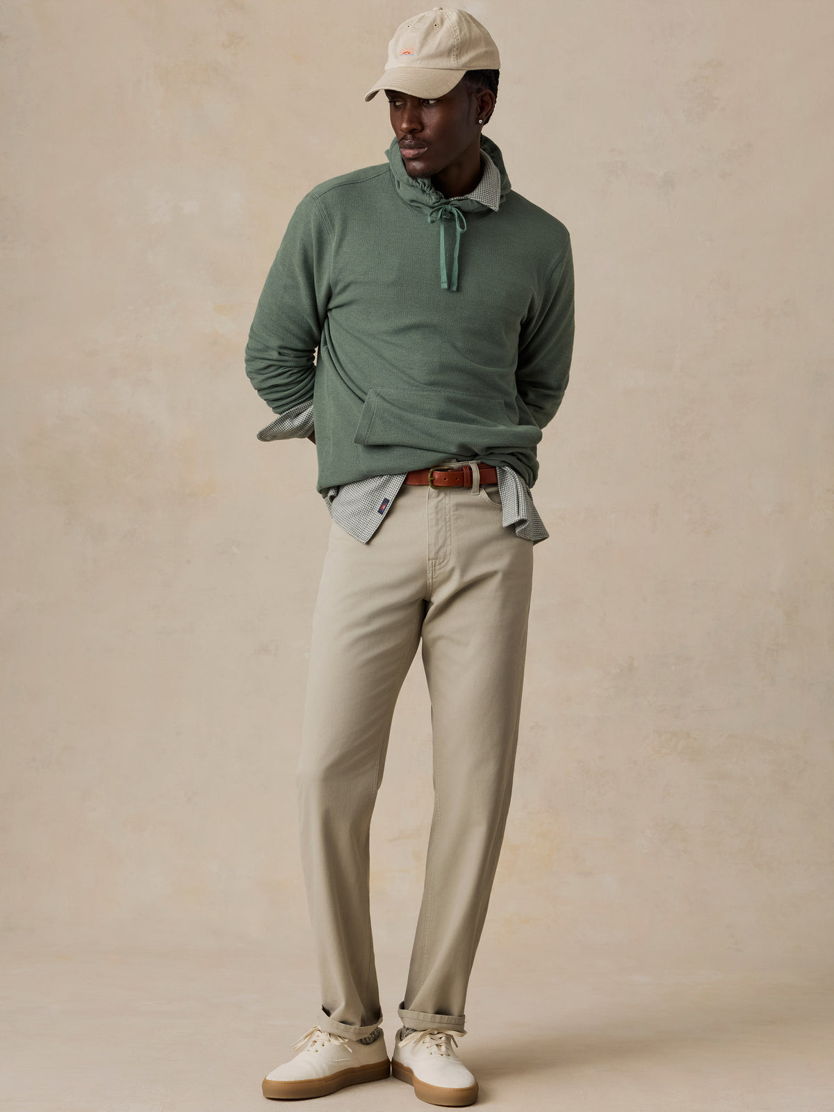 A man modeling a sage green long-sleeve knit polo sweater with a tie-neck detail, styled with beige trousers, a light blue collared shirt, a brown leather belt, and a cream baseball cap against a textured beige studio backdrop., image 2 of 6