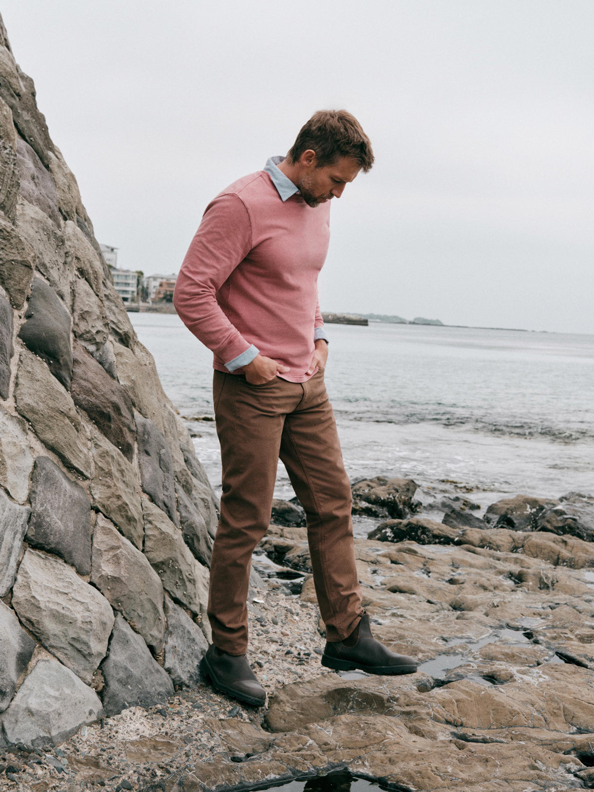 A man wearing a dusty pink long-sleeve sweater, brown trousers, and dark boots stands on a rocky shoreline next to a stone wall, looking down toward the rocks., image 1 of 7