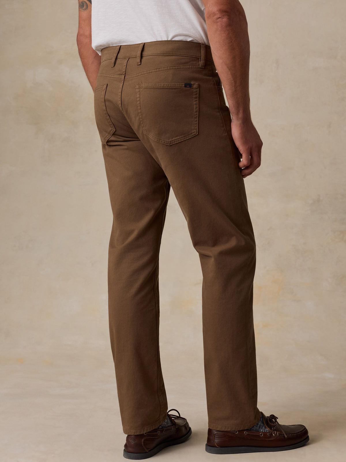 A man shown from the back wearing brown chino pants, a white t-shirt, and brown leather boat shoes, standing against a textured beige studio backdrop., image 6 of 7