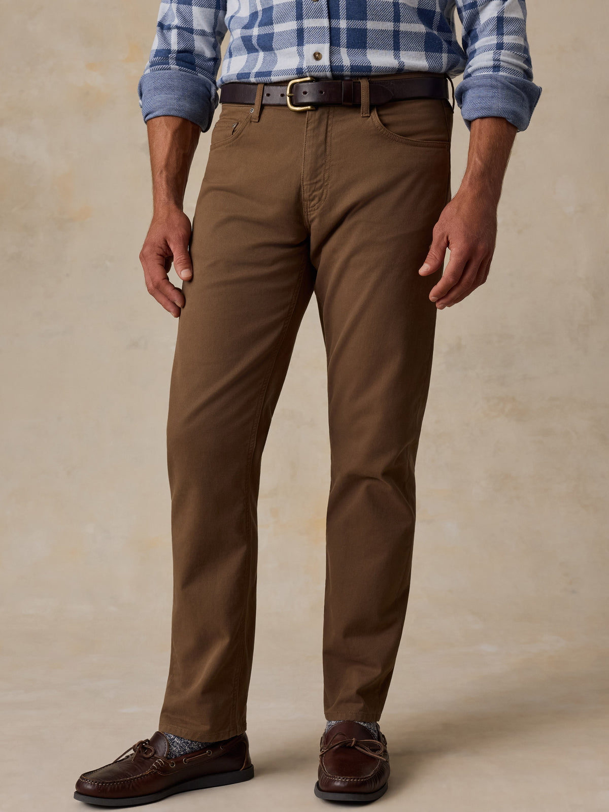 A man wearing brown chino pants, a blue and white plaid shirt, a dark brown belt, and brown leather loafers, standing against a textured beige studio backdrop., image 2 of 7