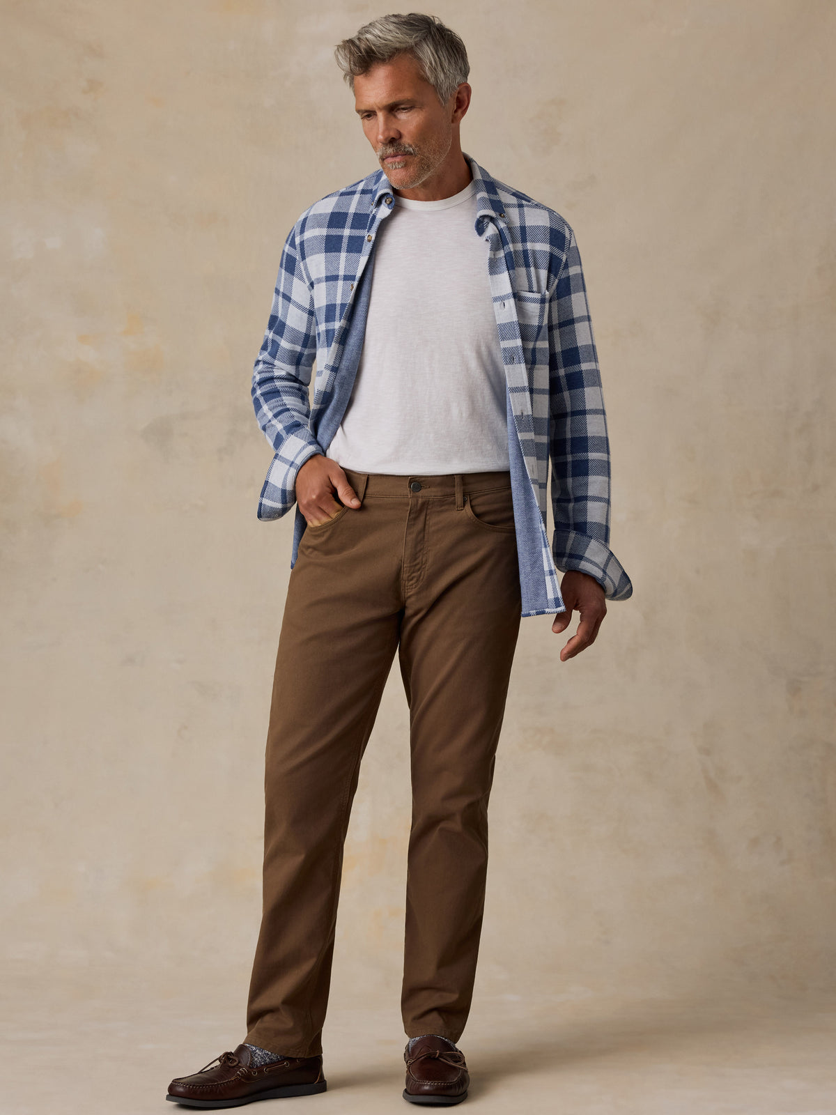 A man modeling a blue and white plaid button-down shirt over a white t-shirt, paired with brown trousers and dark brown leather shoes, standing against a textured beige studio backdrop., image 5 of 7