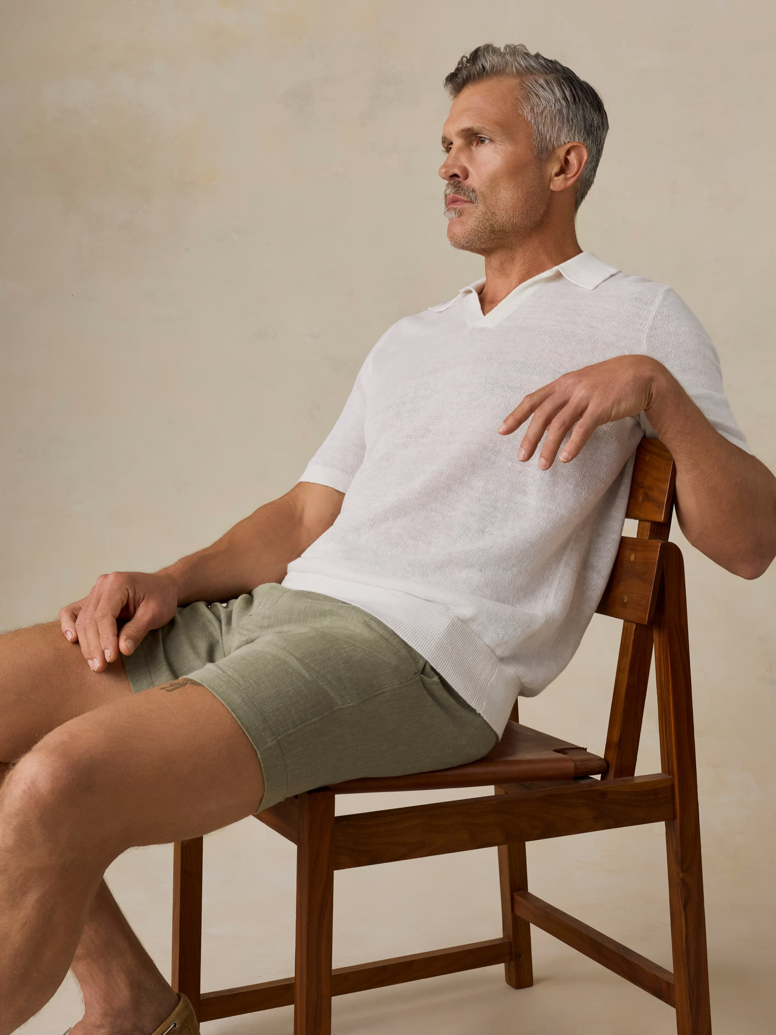Man sitting on a wooden chair wearing a white polo shirt and green linen shorts against a beige background