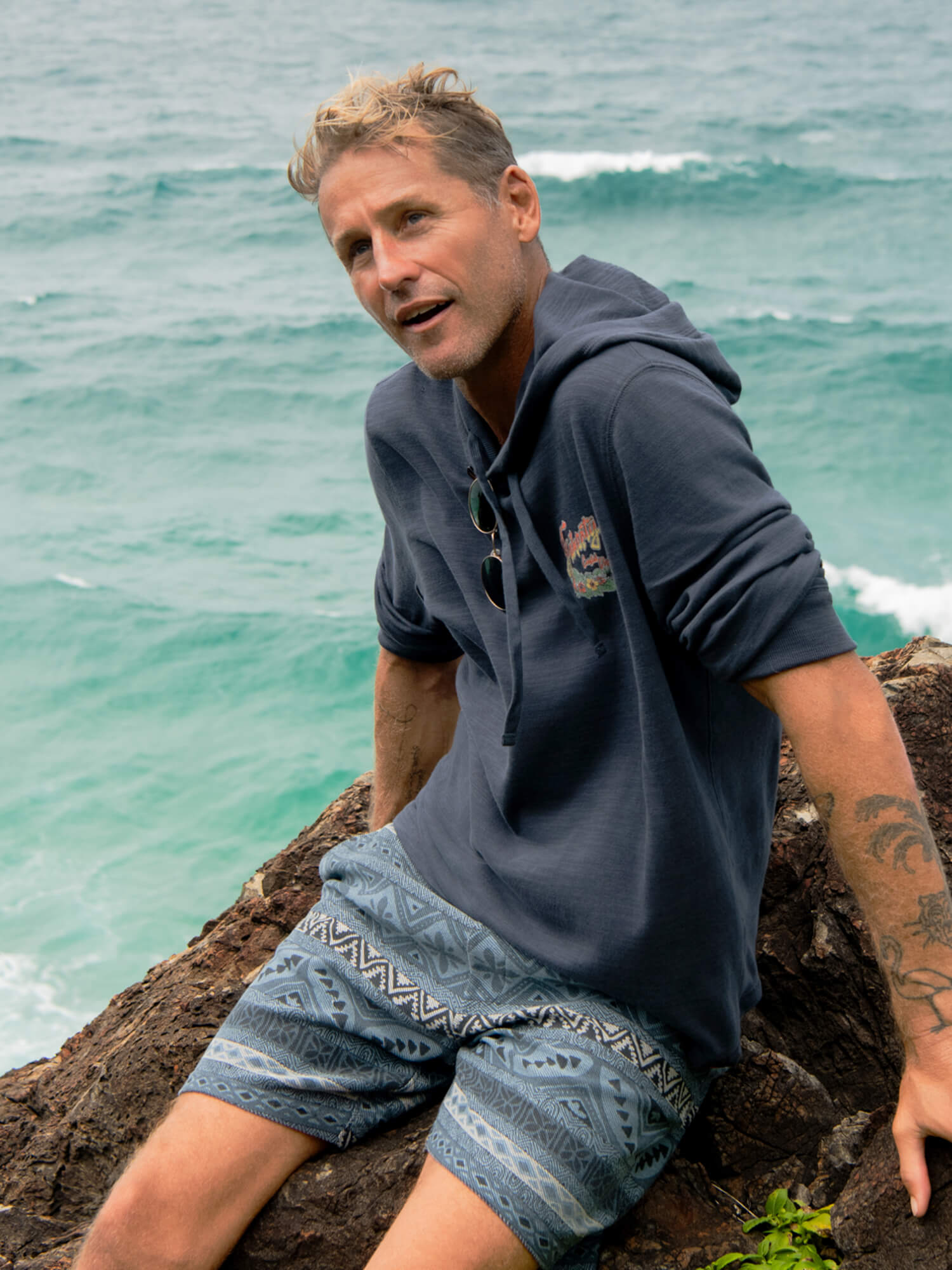 Man wearing printed shorts and a navy hoodie on a rock overlooking the ocean