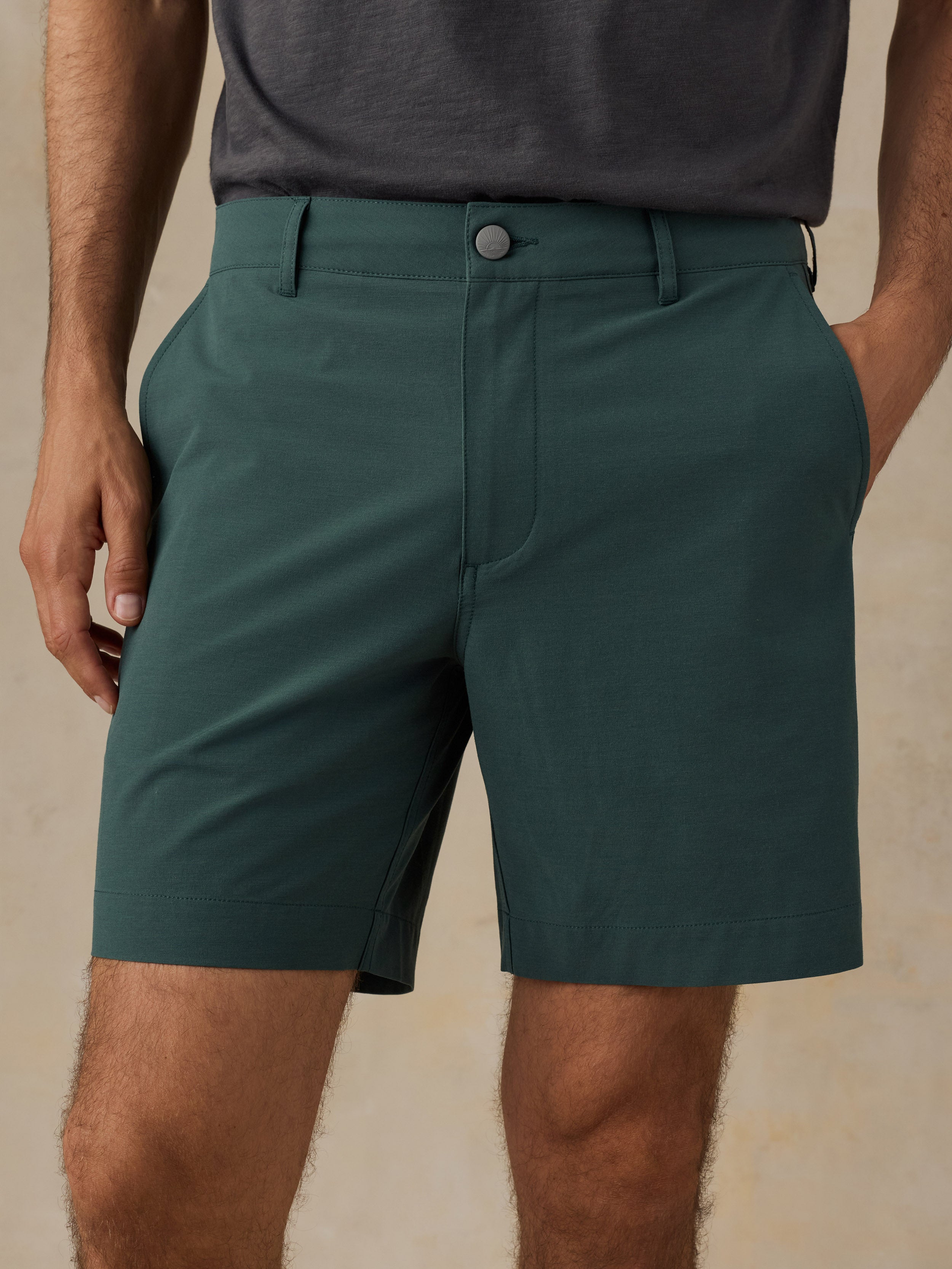 A close-up of a person wearing dark teal-green tailored shorts with a button closure and belt loops, paired with a dark gray shirt against a warm beige textured background.