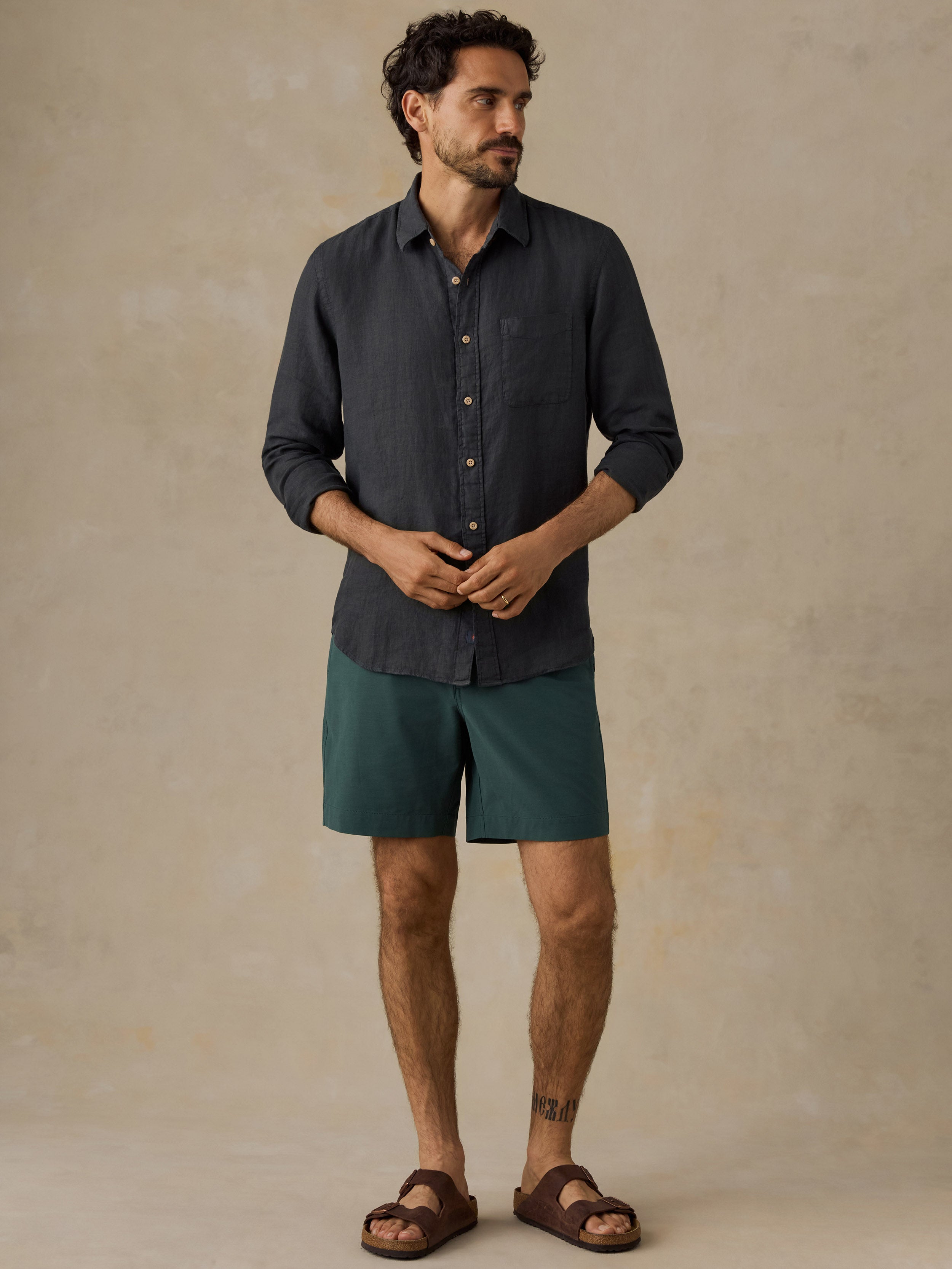 A man wearing a dark charcoal gray long-sleeve button-down shirt, teal green shorts, and brown leather slide sandals, standing against a textured beige studio backdrop.
