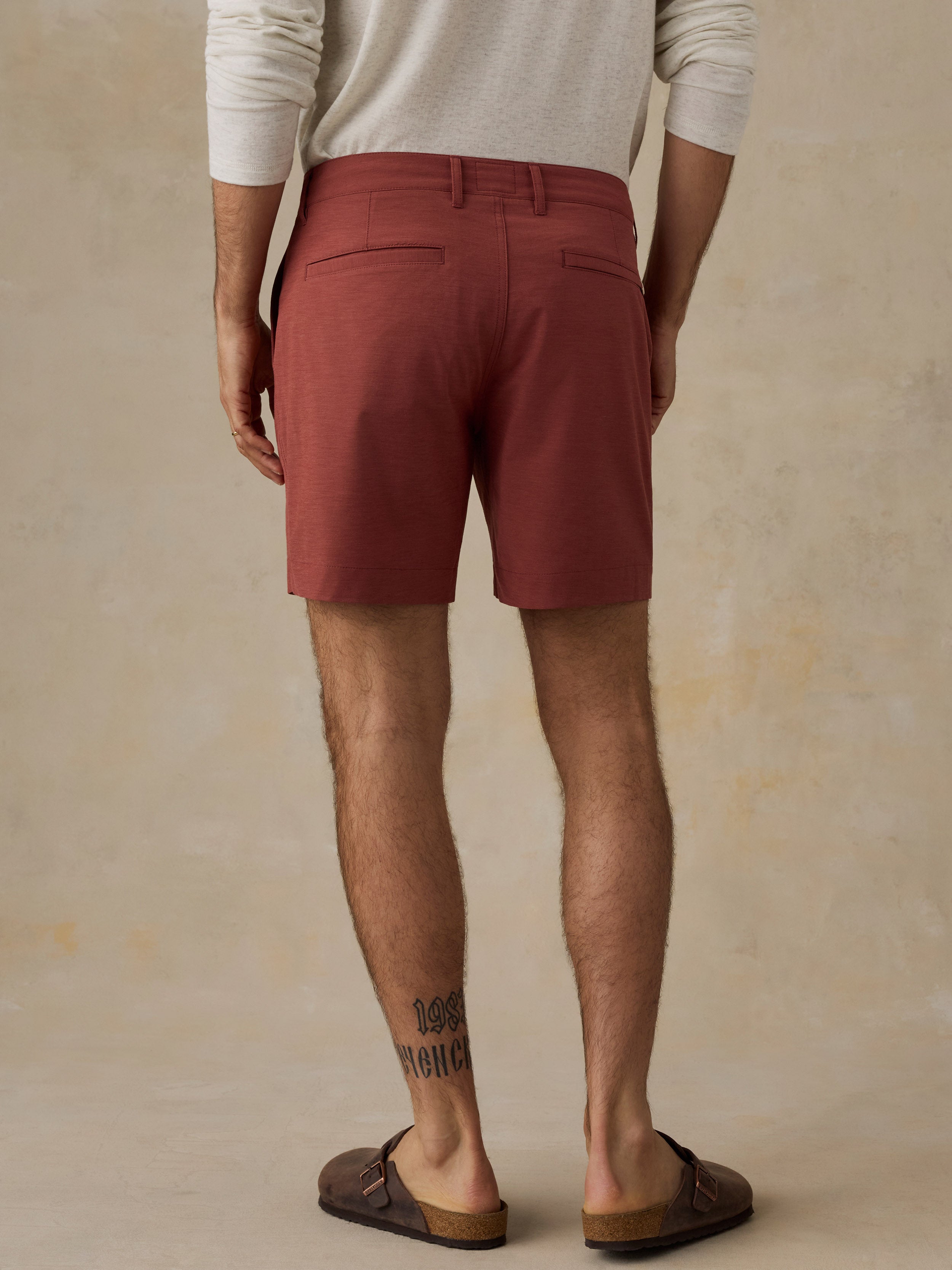 A man viewed from the back wearing rust-red chino shorts, a cream long-sleeve shirt, and brown leather slide sandals against a textured beige studio backdrop.