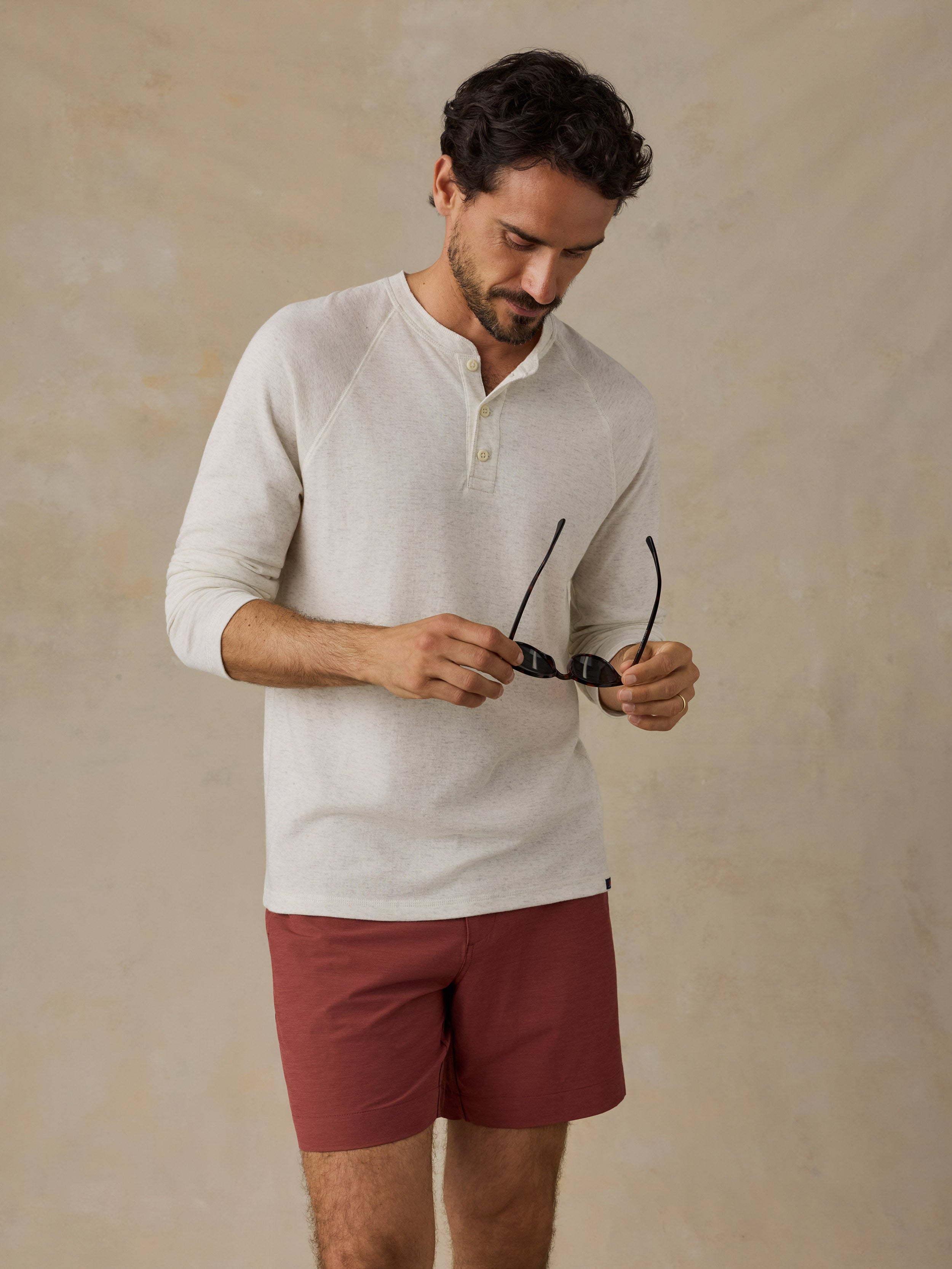 A man wearing a light beige long-sleeve henley shirt and rust-colored shorts, holding a pair of black sunglasses against a textured beige background.
