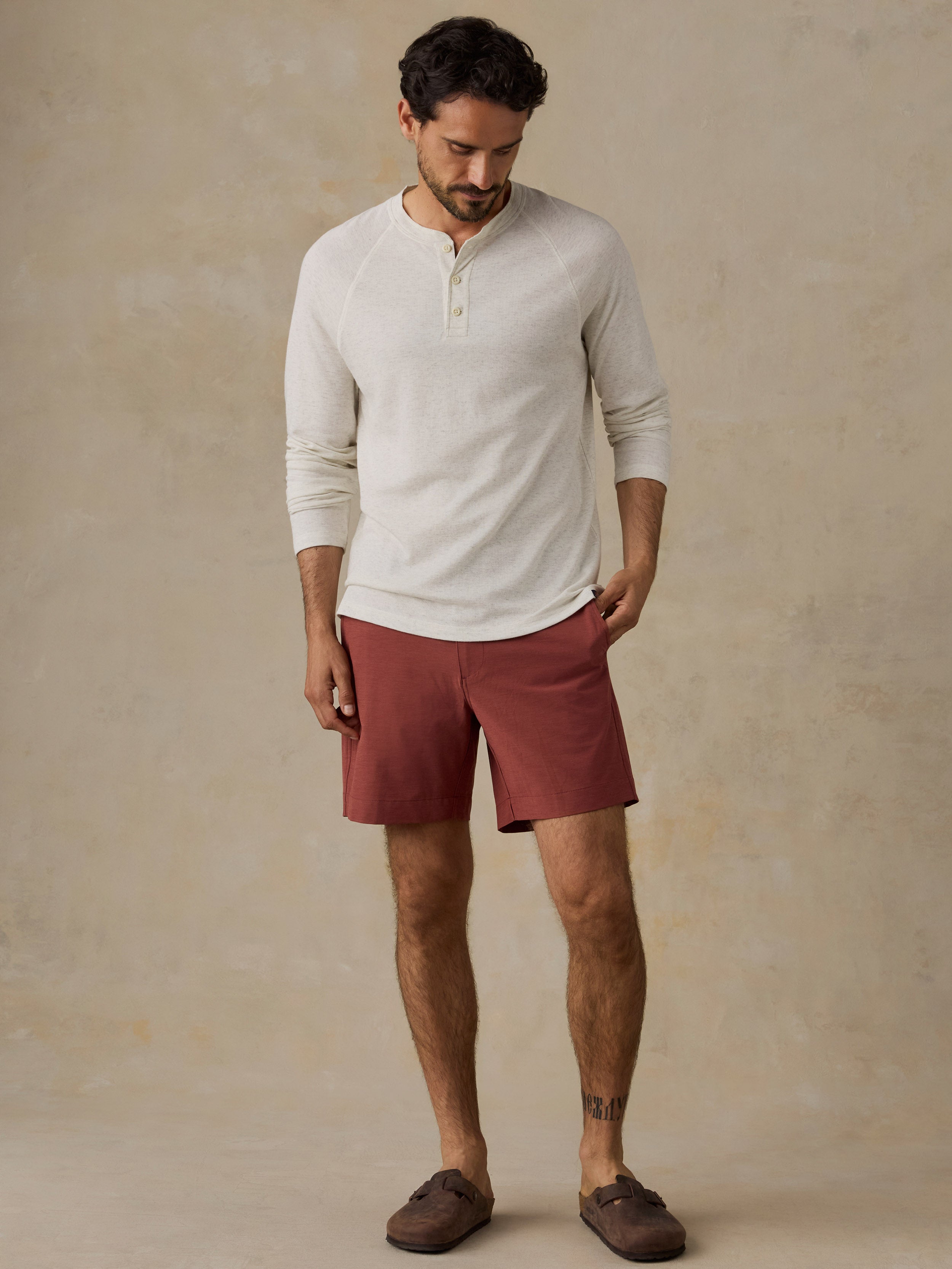 A man wearing a cream-colored long-sleeve henley shirt and rust-red shorts, standing against a textured beige studio backdrop.