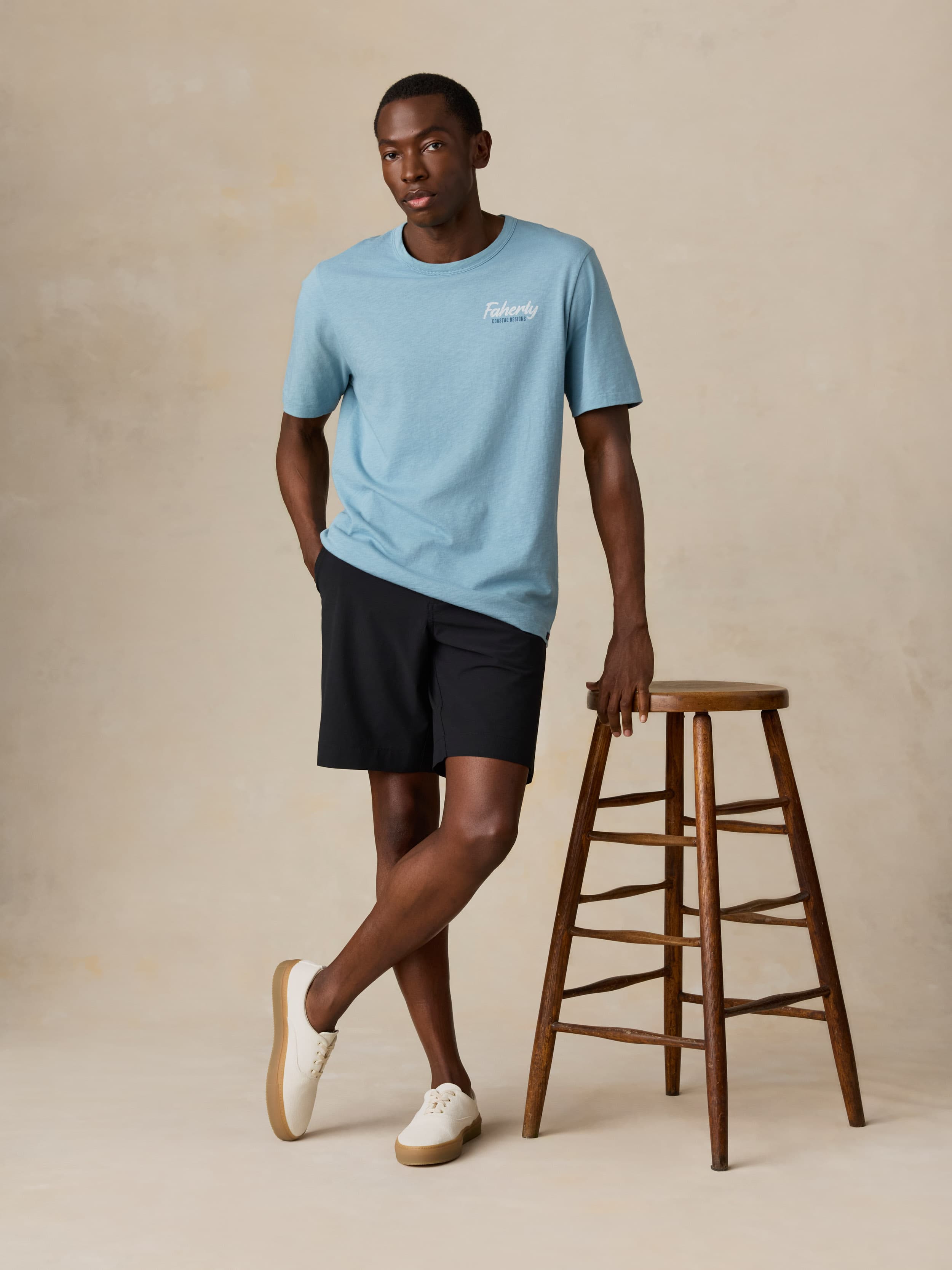 Man wearing a light blue t-shirt and black shorts standing next to a wooden stool on a beige background