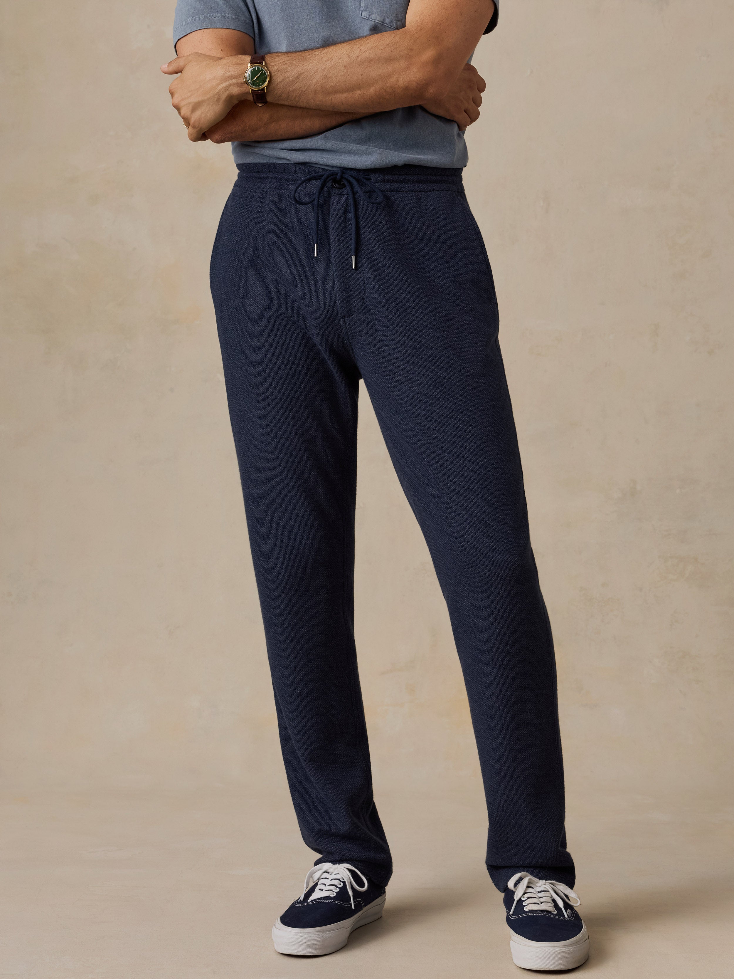 A man wearing dark navy drawstring sweatpants, a blue-gray t-shirt, and navy sneakers, standing against a textured beige studio background.