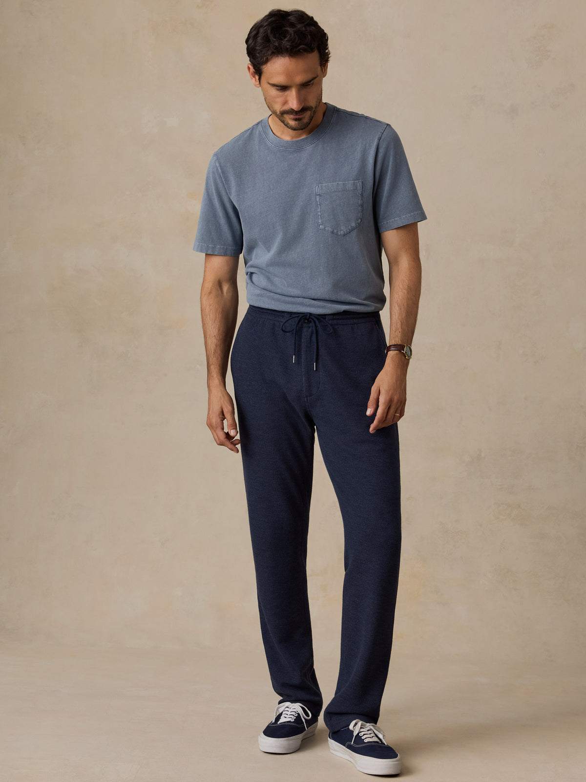 A man wearing a slate blue crewneck pocket t-shirt, navy blue drawstring sweatpants, and navy sneakers, standing against a textured beige studio backdrop., image 2 of 6