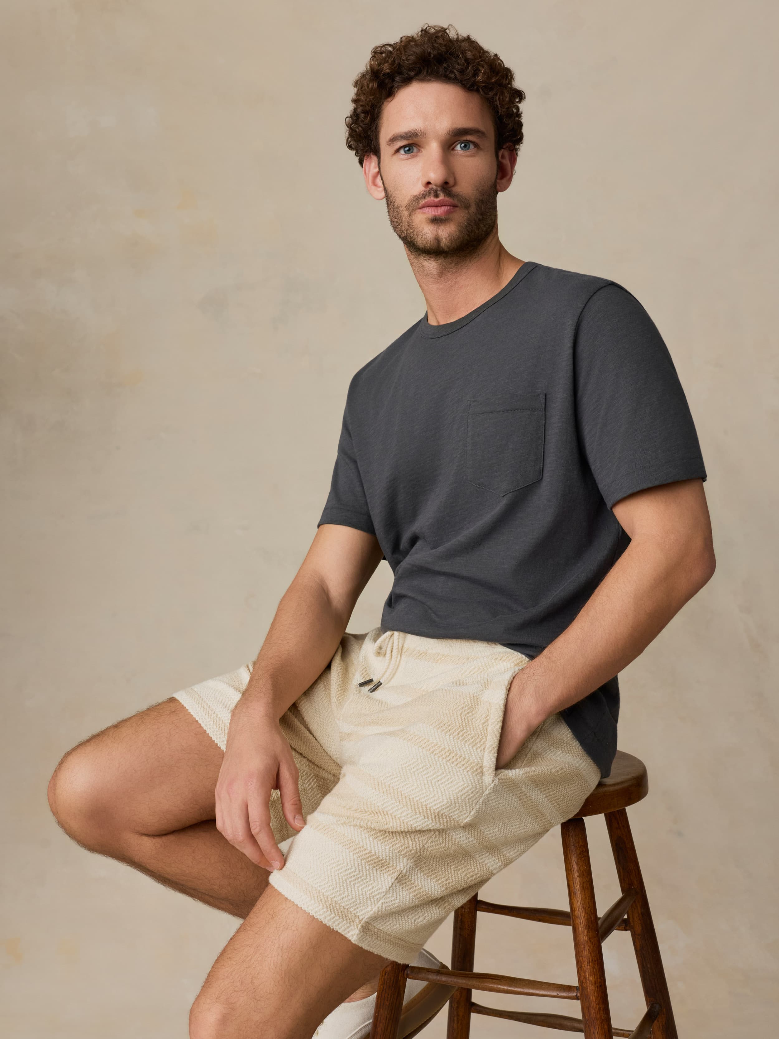 Man sitting on a wooden stool wearing a dark gray t-shirt and beige shorts against a beige background