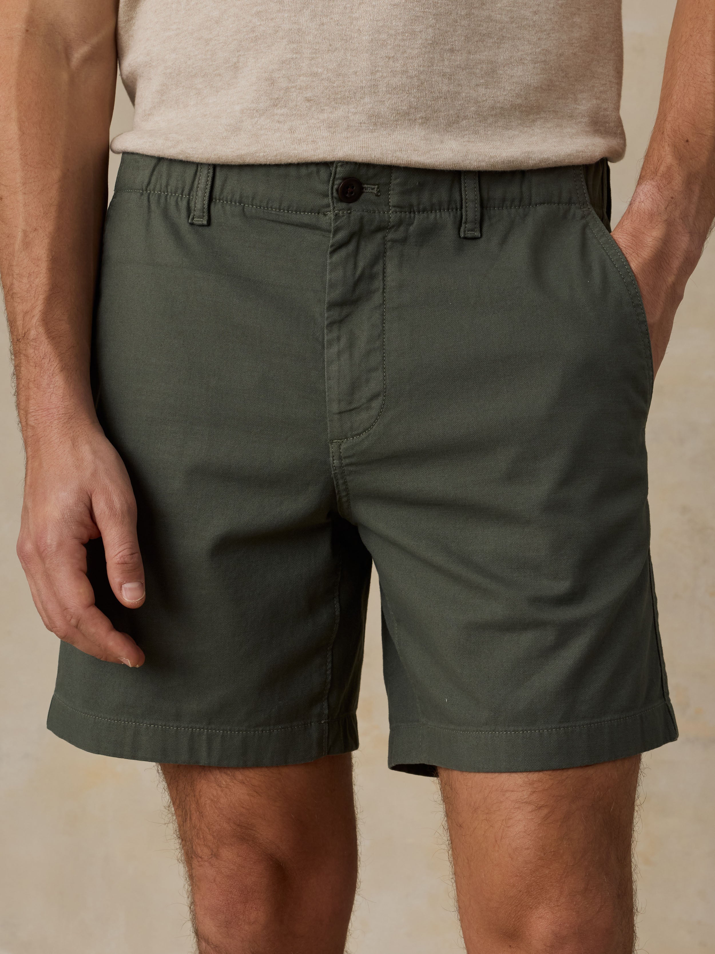 Close-up of a man wearing dark olive green chino shorts with a beige t-shirt against a beige background