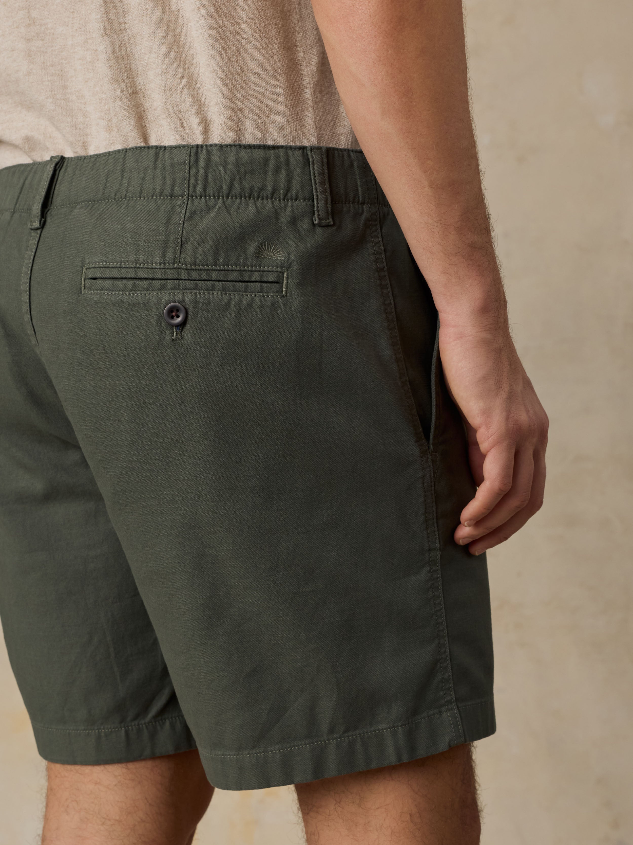 Close-up of a man wearing dark olive green chino shorts with an embroidered Faherty logo above the pocket and a beige t-shirt against a beige background