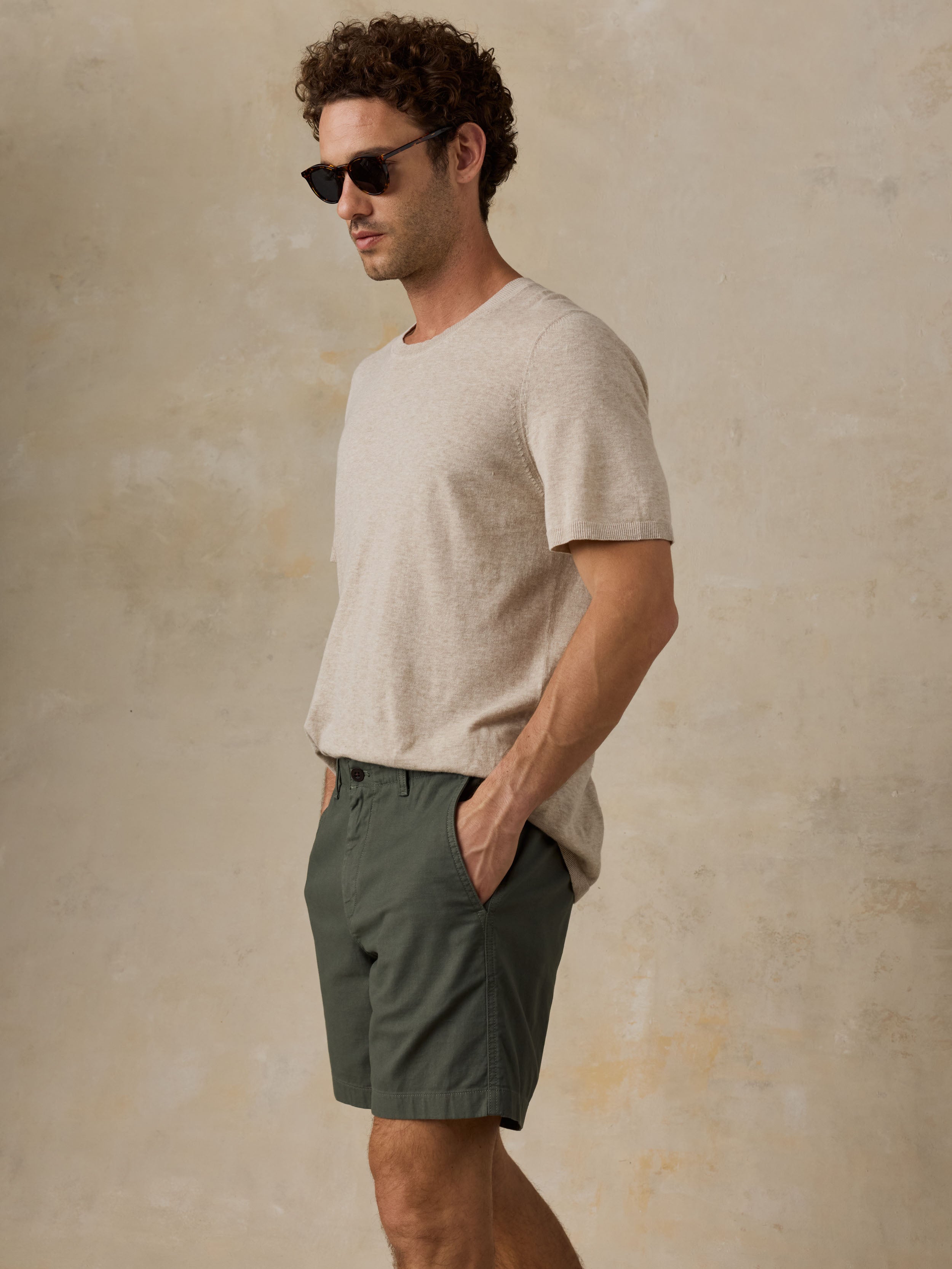 Man wearing dark olive green chino shorts with a beige t-shirt, sandals, and sunglasses against a beige background